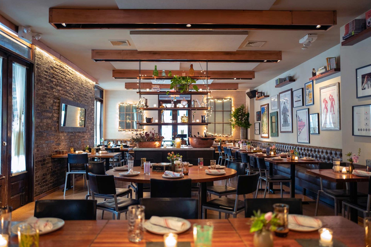 A cozy, modern restaurant with wooden beams, string lights, and framed art on the walls. Tables are set for dining with candles and glassware, and shelves with plants divide the space.