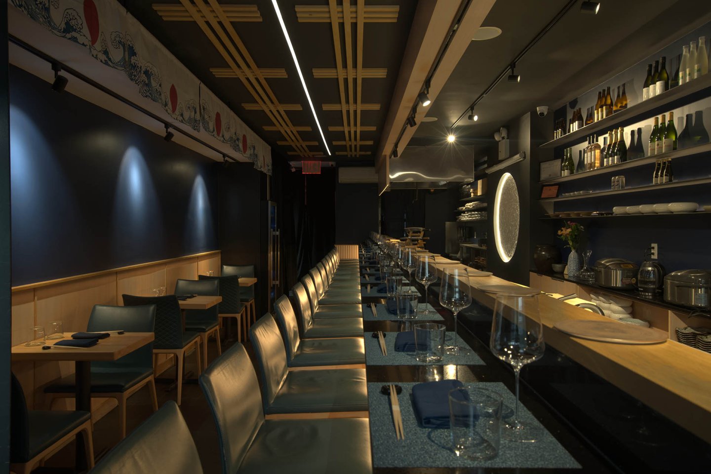A modern, dimly lit restaurant interior with a long bar, high chairs, neatly set tables, and shelves holding bottles and glassware. The atmosphere is elegant and inviting.
