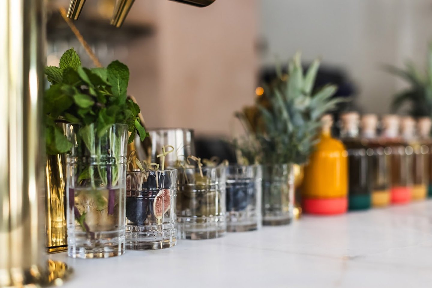 A row of clear glasses filled with fresh herbs, fruits, and garnishes sits on a white counter, with bottles of colorful liquids blurred in the background.