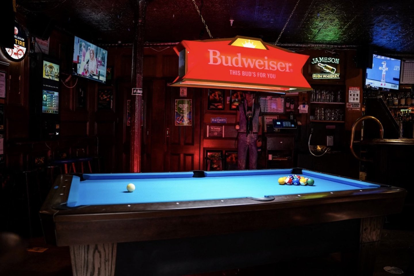 A dimly lit bar with a blue pool table, billiard balls arranged for a break, and a red Budweiser light hanging overhead. TV screens, posters, and a stocked bar are visible in the background.