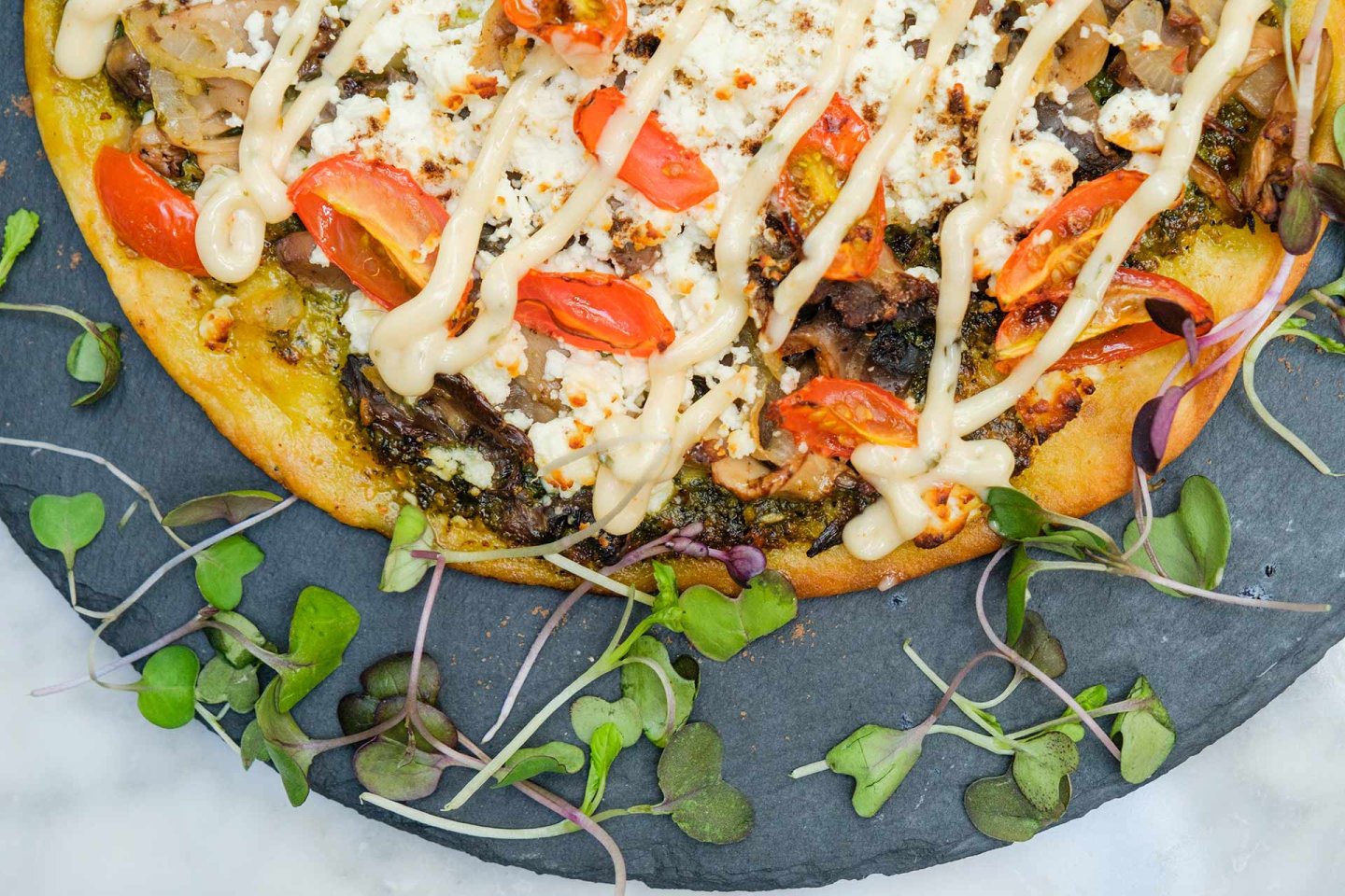 A close-up of a gourmet pizza topped with tomatoes, onions, cheese, and creamy sauce, surrounded by microgreens on a slate serving tray.