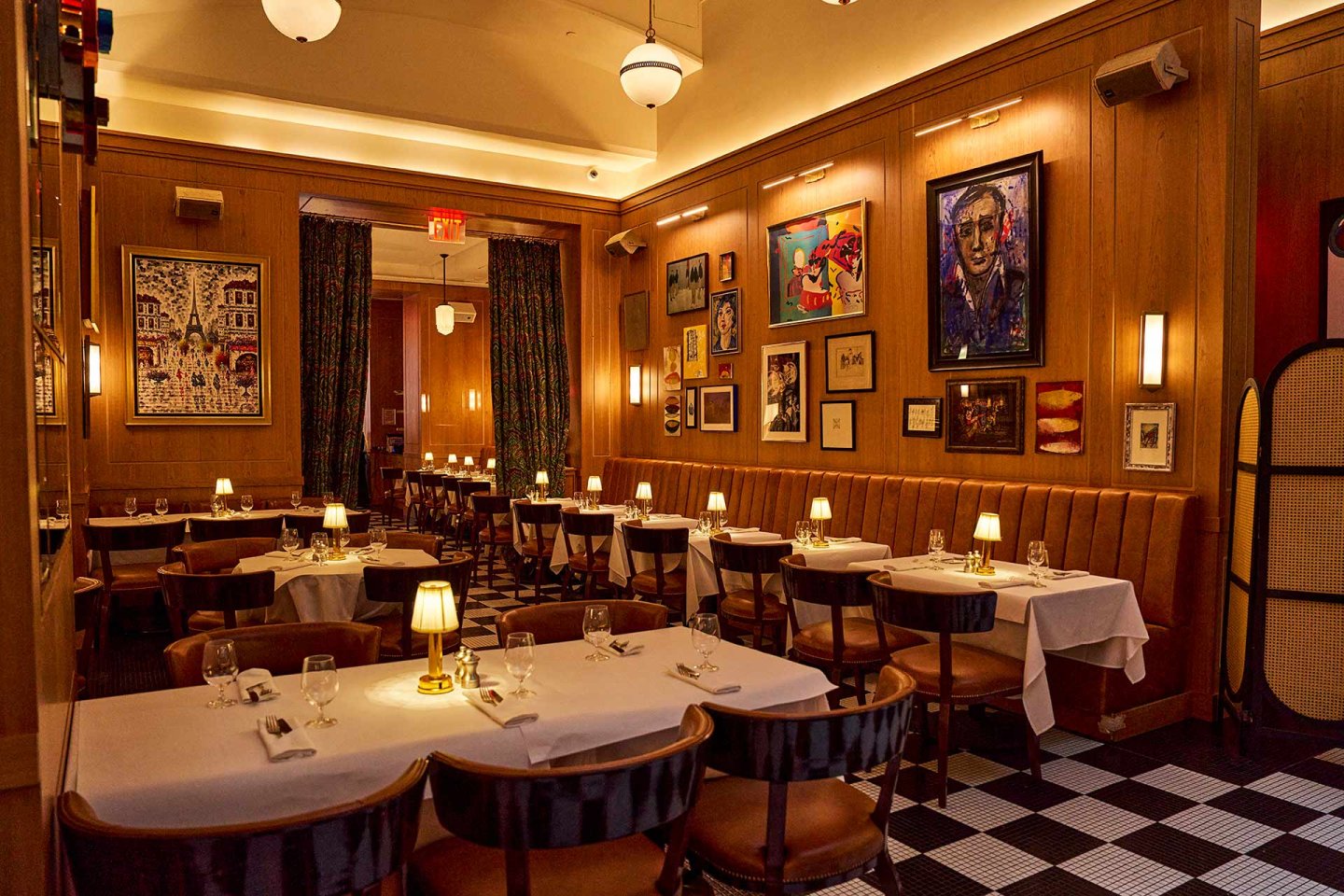 A warmly lit restaurant with checkered floors, tan leather banquettes, and round tables set with white cloths. Colorful artwork covers the wood-paneled walls, and small lamps provide a cozy ambiance.