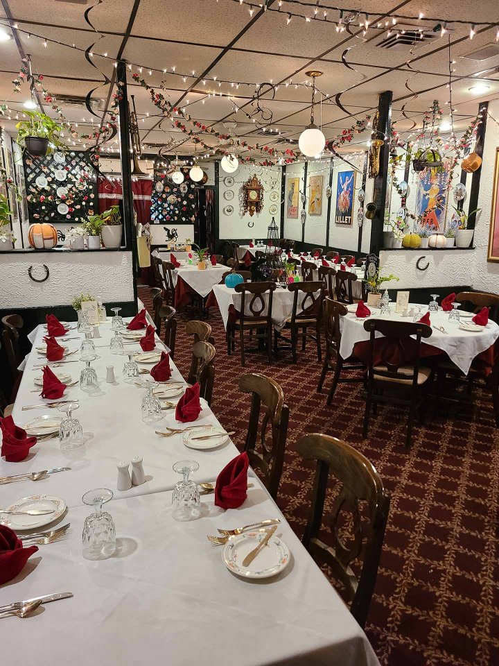 A cozy restaurant dining room with long tables set for a meal, featuring white tablecloths, red napkins, wooden chairs, string lights, and walls decorated with framed art and various hanging plants.