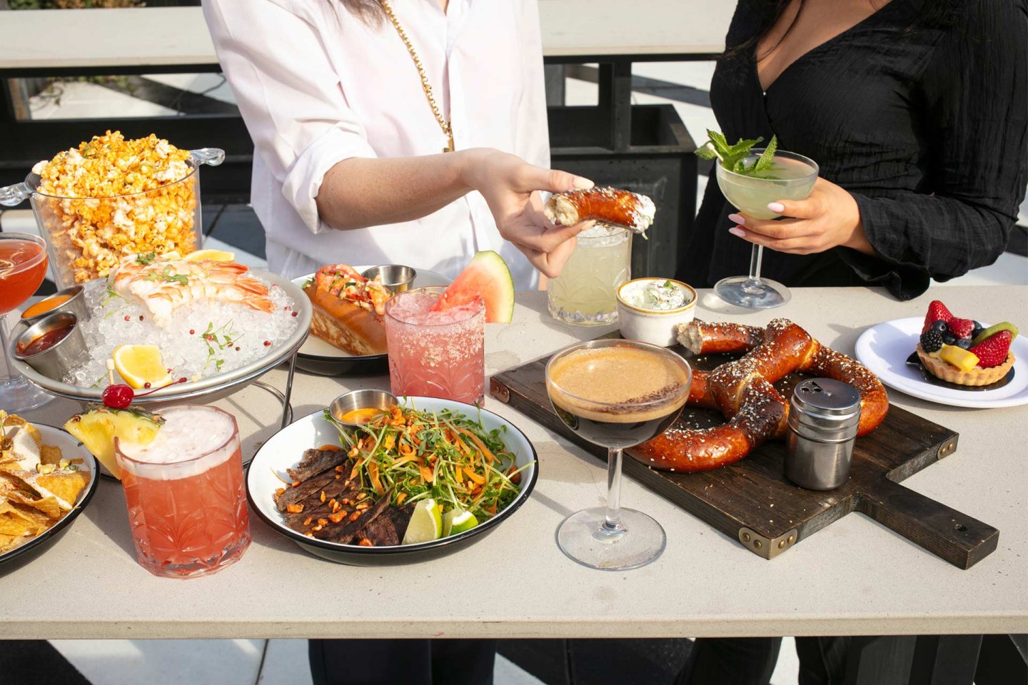Two women enjoy a table full of food and drinks, including pretzels, shrimp cocktail, popcorn, cocktails, steak salad, a hot dog, dips, and fruit tarts, all arranged for a casual outdoor meal.