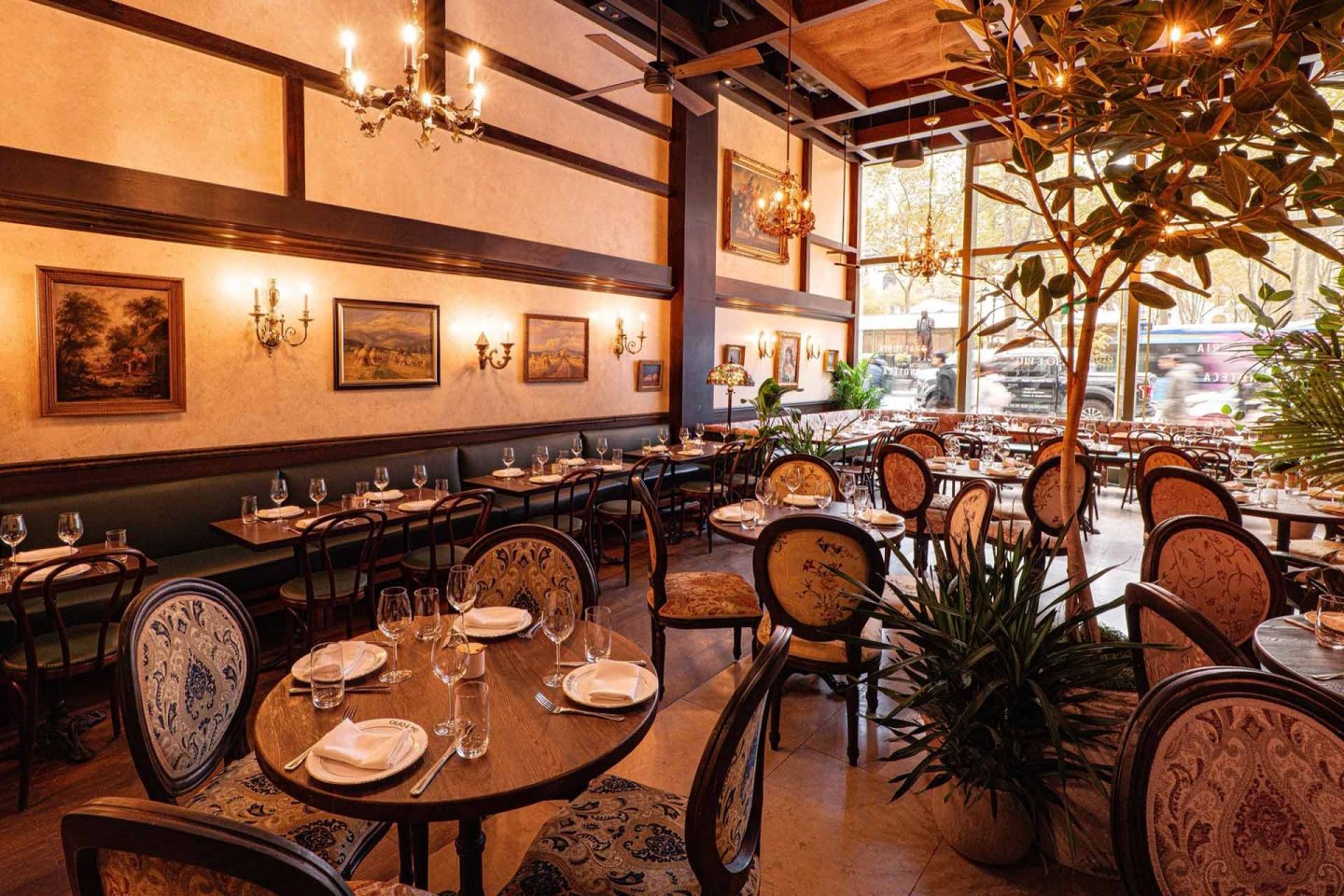 Elegant restaurant interior with round tables set for dining, upholstered chairs, classic chandeliers, framed paintings on cream walls, large windows letting in natural light, and green plants adding a cozy ambiance.