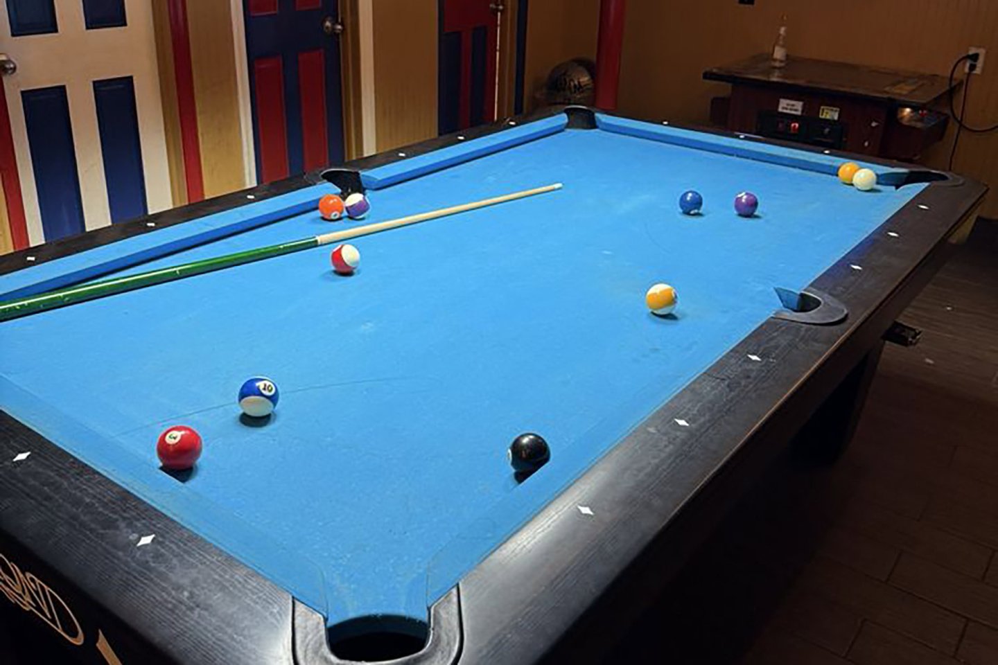A blue-felt pool table with several balls scattered across the surface and a cue stick resting diagonally on the table. The room has red and blue accented doors and a wooden floor.