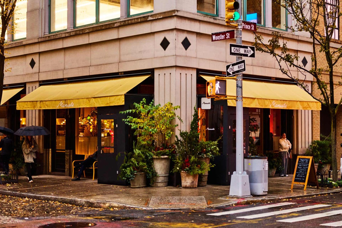 A corner café with yellow awnings and large windows, surrounded by potted plants. People stand near the entrance, and a street sign marks the intersection of Wooster St and Prince St on a rainy day.