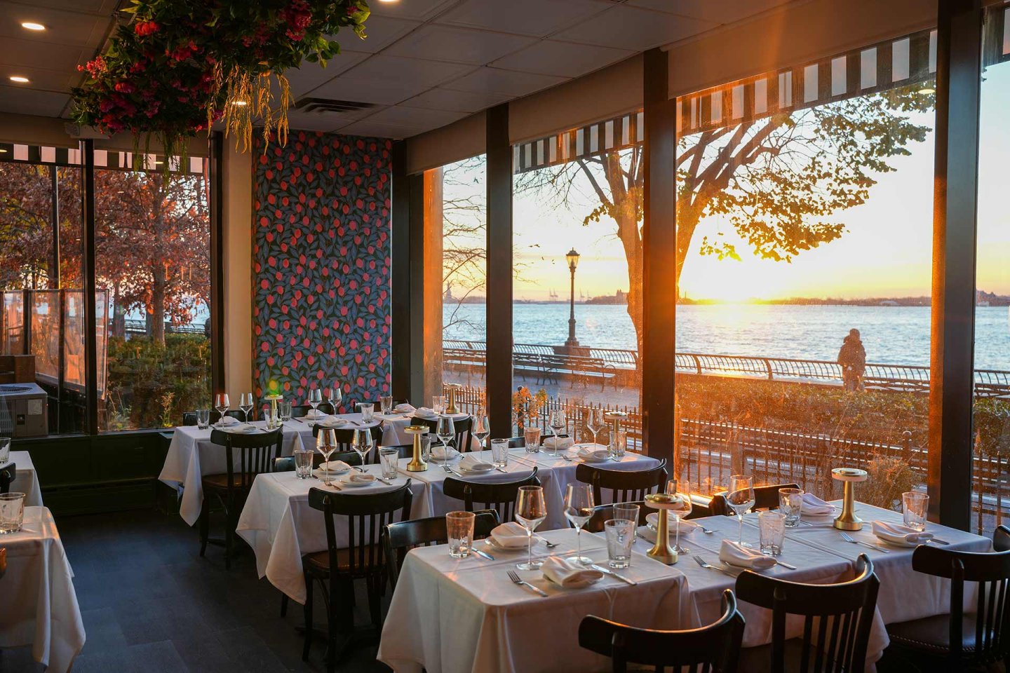Elegant restaurant with tables set for dining, large windows overlooking a waterfront at sunset, casting warm light inside. A person walks along the promenade outside. There are patterned wallpaper and hanging flowers inside.