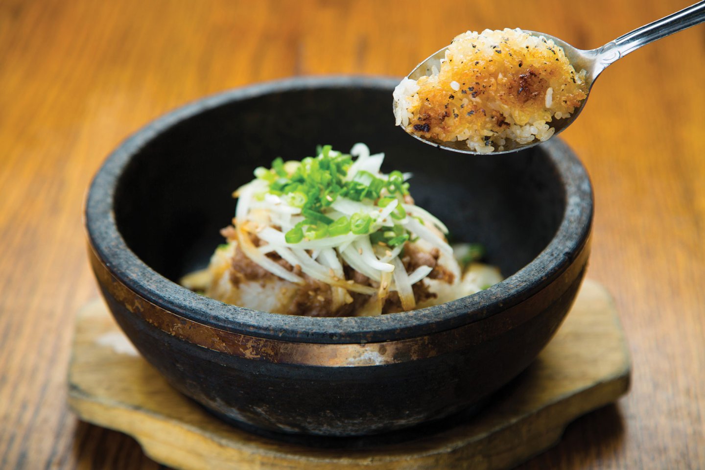 A spoonful of crispy rice is held above a hot stone bowl filled with rice, vegetables, and green onions, set on a wooden table.