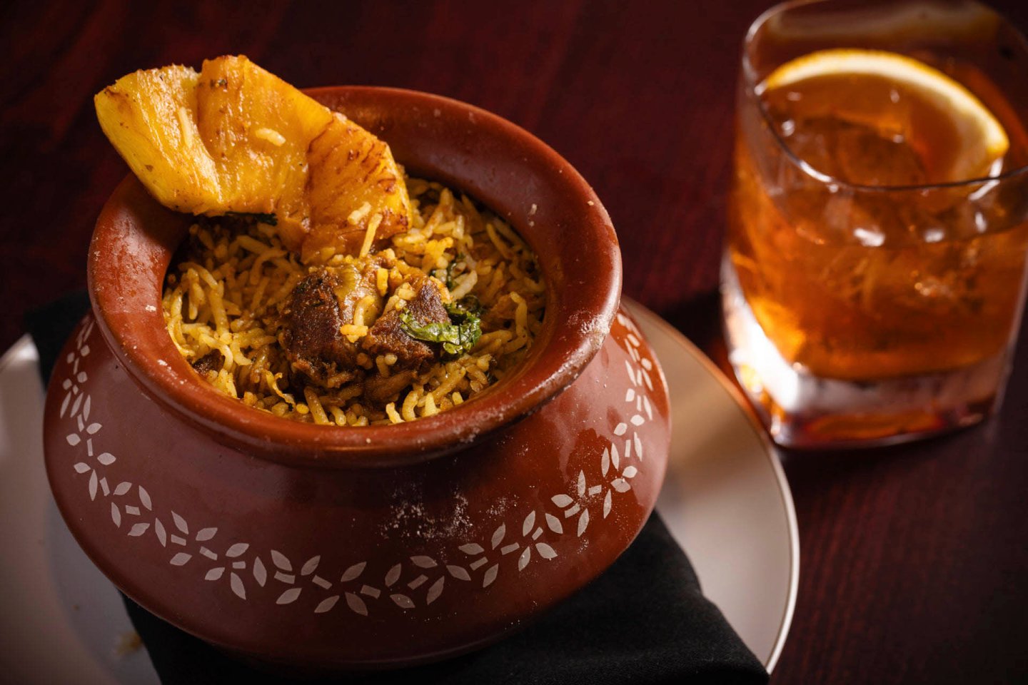 A clay pot filled with fragrant biryani, garnished with a slice of pineapple, sits on a plate with a black cloth. Beside it is a glass of amber-colored drink with an orange slice. The setting is on a dark wooden table.
