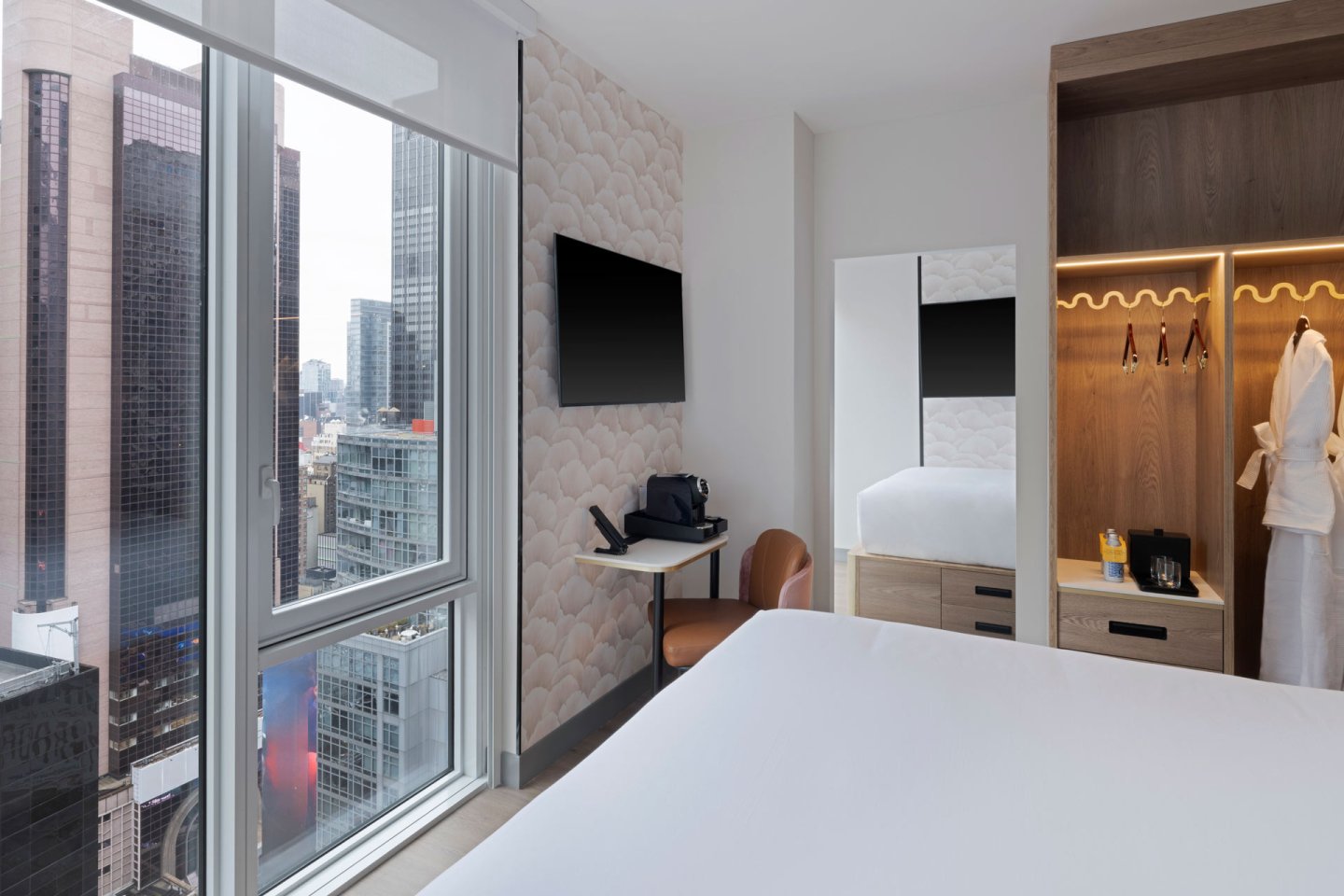A modern hotel room with a large bed, wall-mounted TV, desk, open closet, and city view through floor-to-ceiling windows. A robe hangs in the closet, and tall buildings are visible outside.