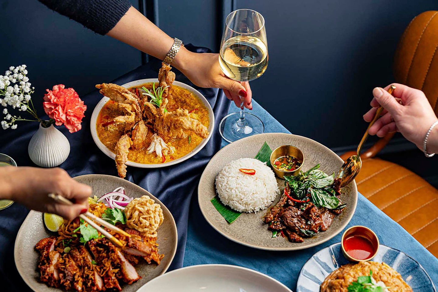 A table with a variety of Asian dishes, including a bowl of crispy fried crab, a plate of rice with beef and greens, and slices of roasted meat with noodles. Hands are reaching for the food and a glass of white wine.