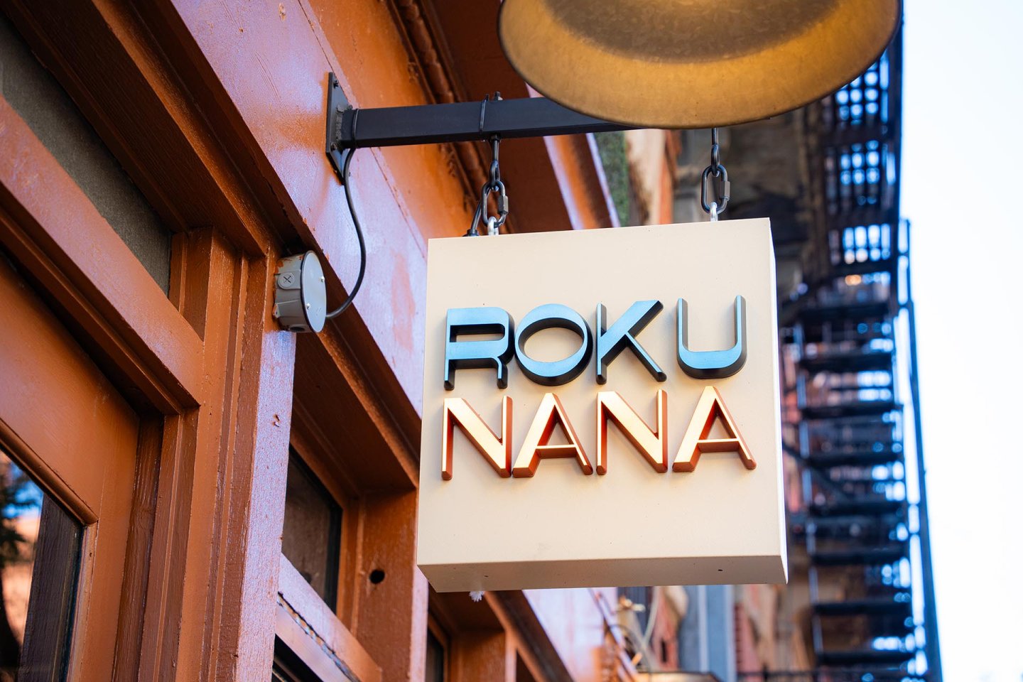 A white sign hanging outside a building reads "ROKU NANA" in blue and orange letters, with part of a brown exterior wall and a metal staircase visible in the background.