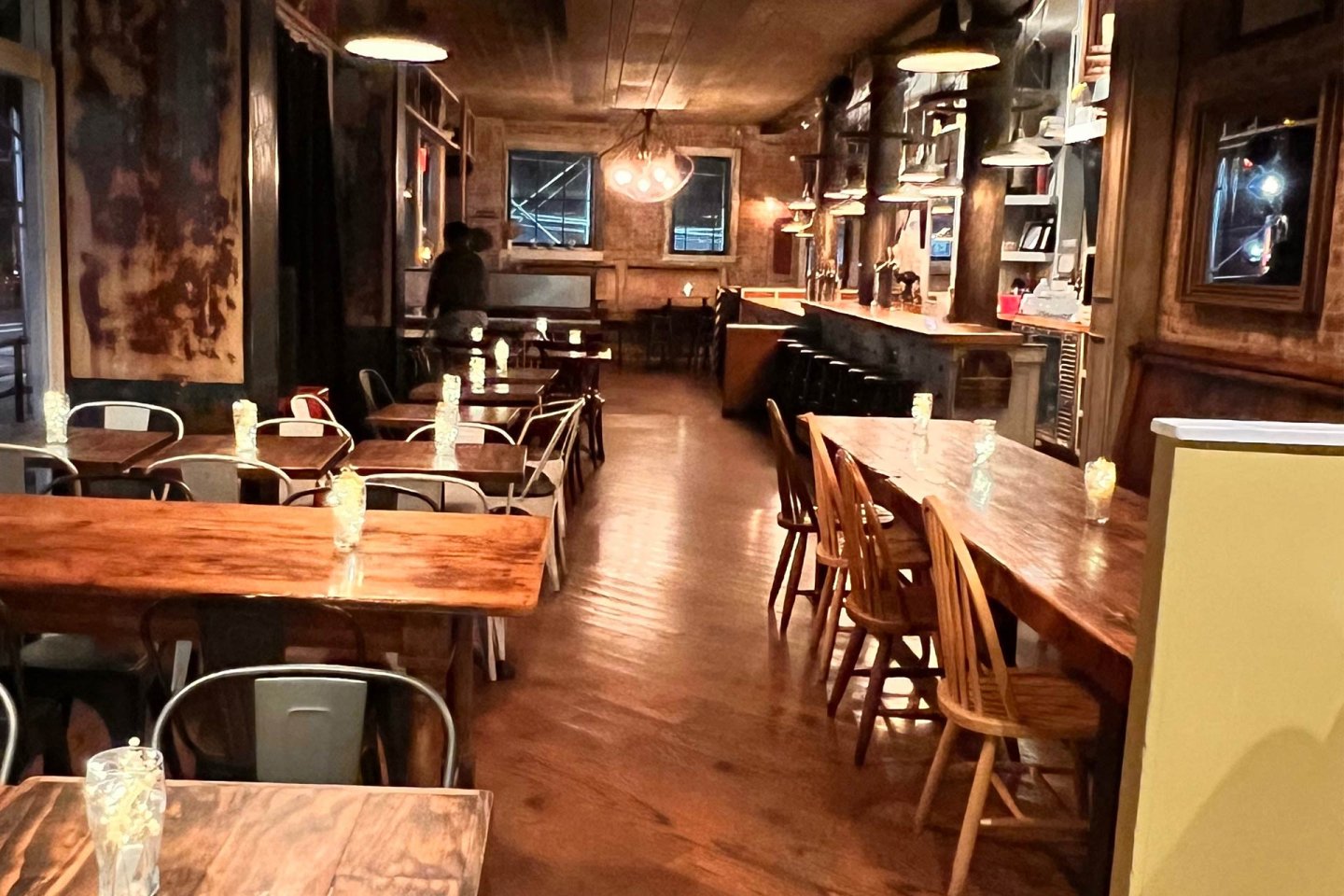 A warmly lit, rustic restaurant interior with wooden tables and chairs, candles on tables, and a bar area in the background. One person stands near the back wall. Large windows show lights from outside.