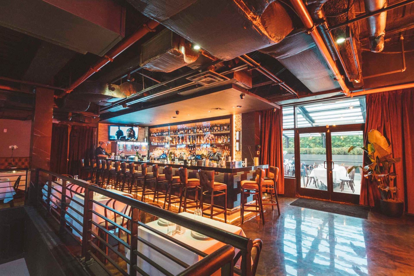 A cozy bar interior with a wooden counter lined with high chairs. Shelves behind the bar are stocked with bottles. Warm lighting enhances the ambiance. Large windows and doors lead to an outdoor seating area.