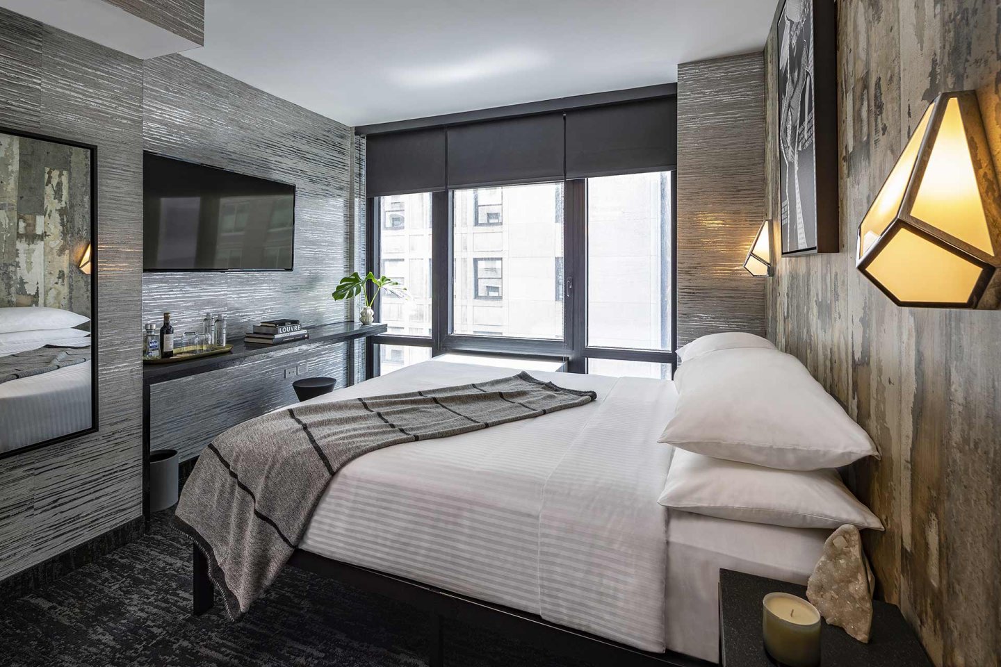 Modern hotel room with a neatly made bed, gray blanket, wall-mounted TV, mirror, desk with a chair, two geometric wall lights, large windows with city views, and contemporary decor.