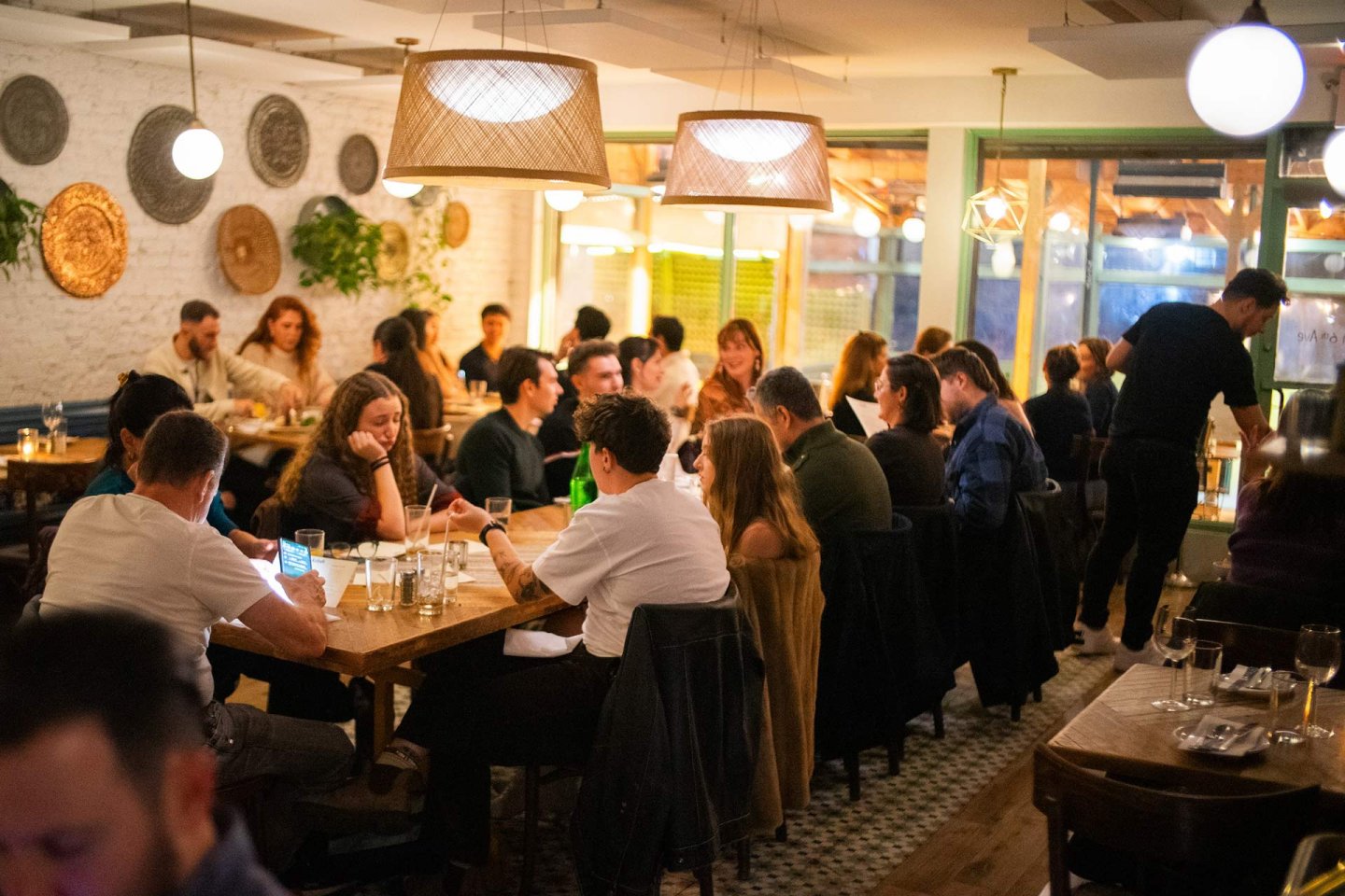 A busy restaurant with people sitting at wooden tables, eating, drinking, and talking. Warm lighting and decorative wall plates create a cozy atmosphere. A server is taking orders from one of the tables.