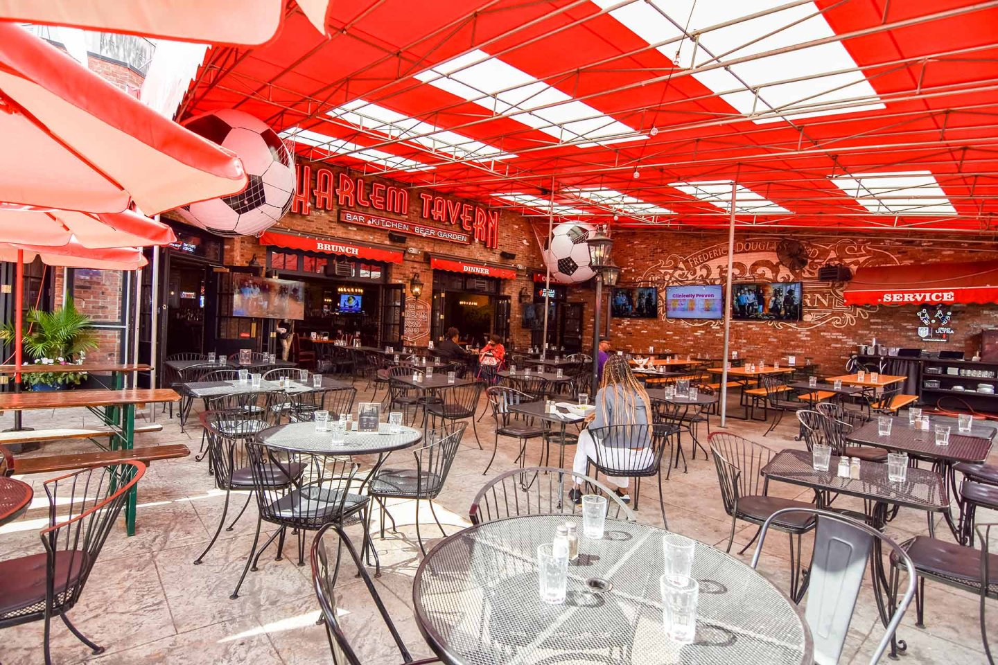 Outdoor seating area at Harlem Tavern, featuring metal tables and chairs under a red canopy, large soccer ball decorations, and brick walls with TVs and signage in the background.