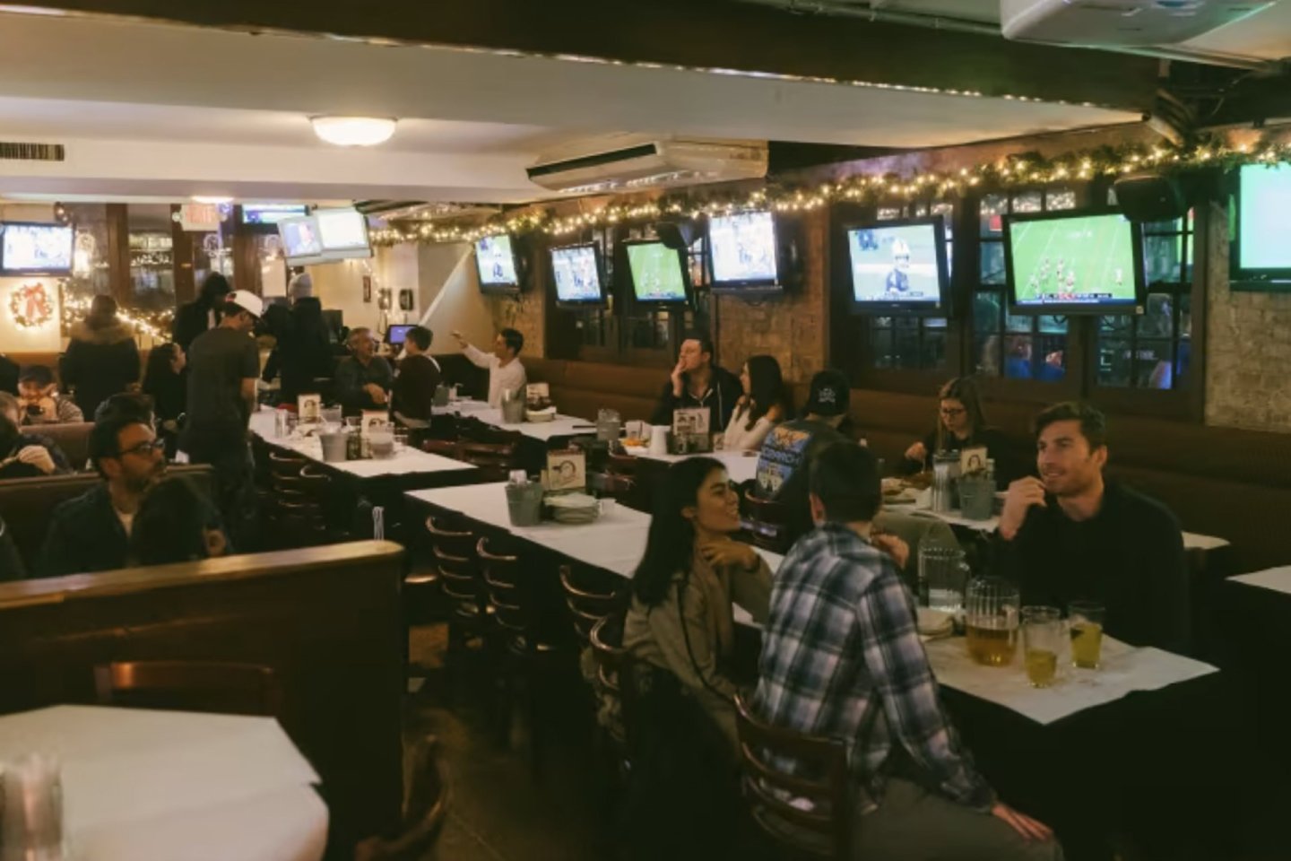 A lively bar with people sitting at tables, drinking beer, and chatting. Multiple TVs displaying sports line the walls. The room is decorated with string lights, creating a warm, inviting atmosphere.