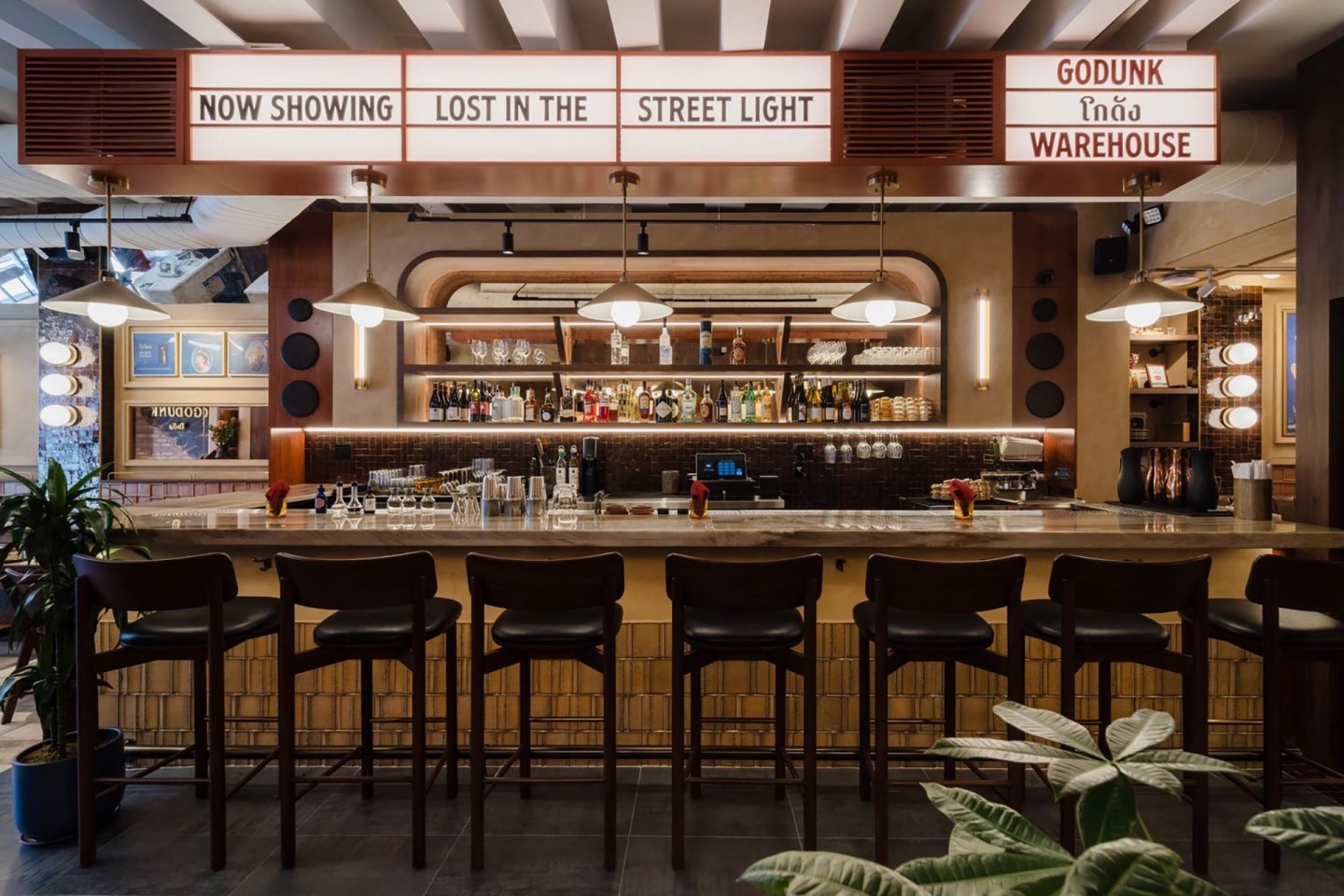 A modern bar with six high chairs, a well-stocked backlit shelf of bottles, a counter with glassware, and overhead marquee signs reading “Now Showing Lost in the Street Light” and “Godunk Tína Warehouse.”.