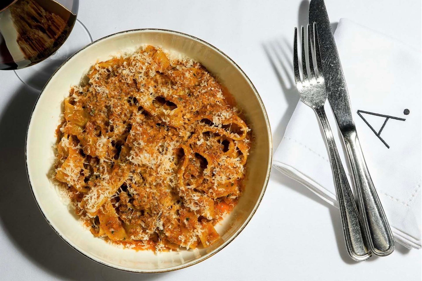 A bowl of pasta with meat sauce and grated cheese is on a white tablecloth next to a glass of red wine and a napkin with a fork and knife.
