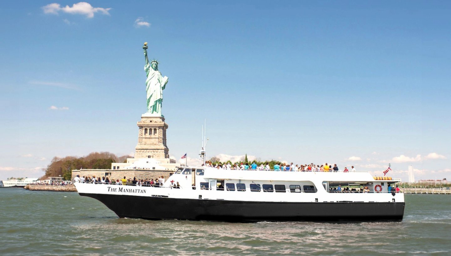 A white and black Liberty Cruise sightseeing boat filled with passengers sailing on the water in front of the Statue of Liberty under a clear blue sky.