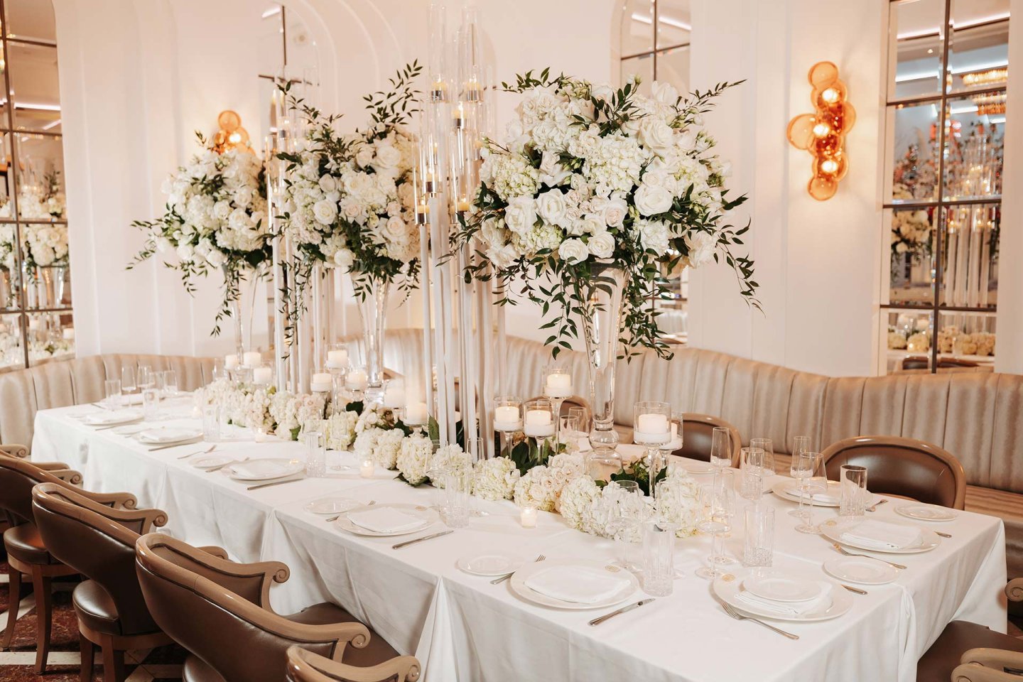 Elegant dining table set for a formal event, decorated with tall glass vases of white flowers and greenery, surrounded by beige chairs, white linens, and softly lit candles in a luxurious room.