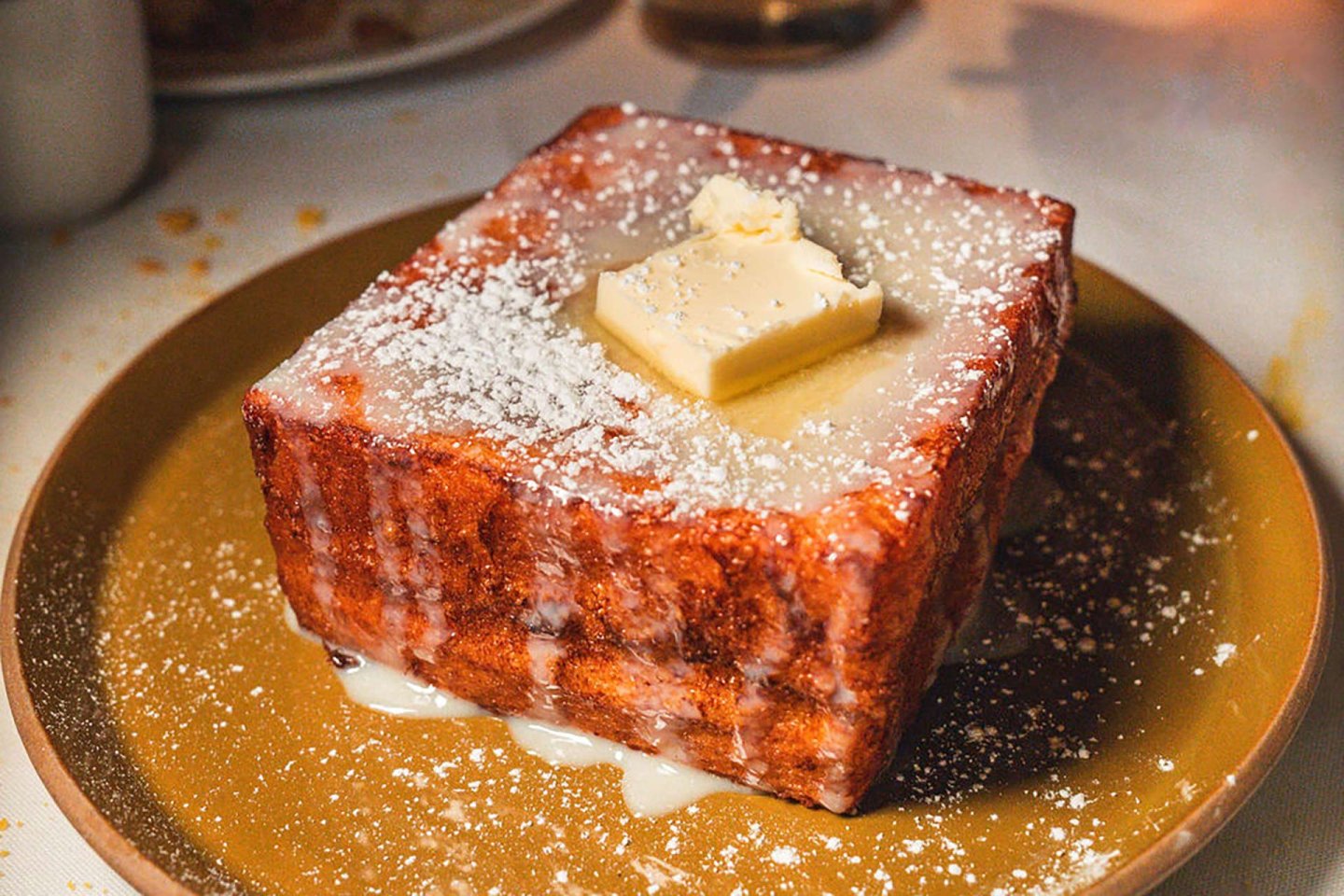 A thick, square piece of French toast is topped with a pat of butter and powdered sugar, served on a brown plate with a drizzle of syrup or glaze.