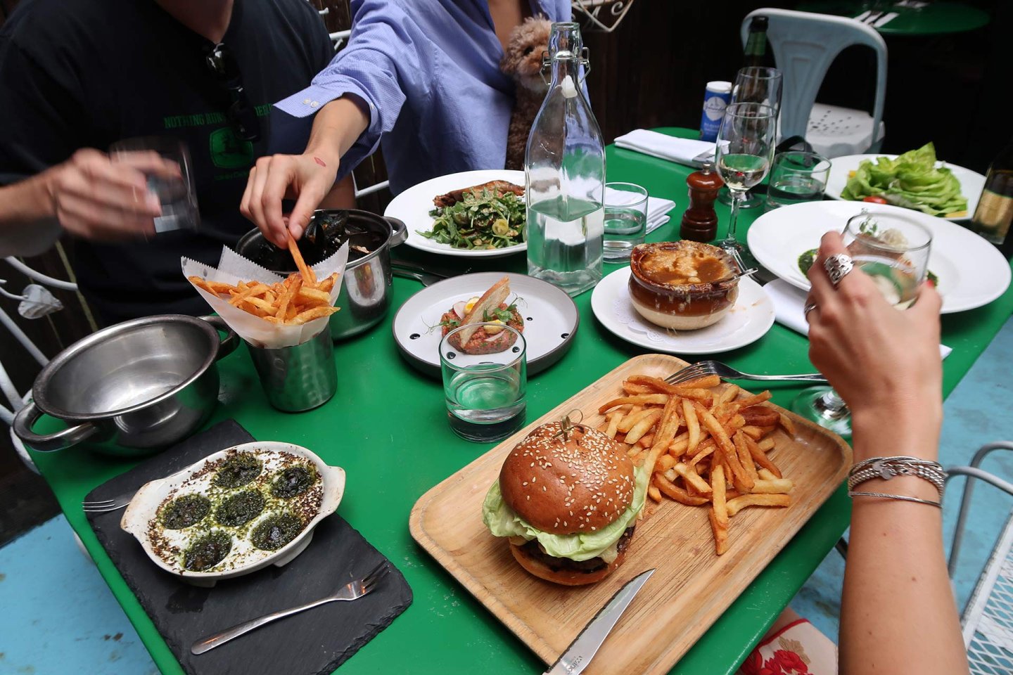 A group of people share a meal at a green table with burgers, fries, escargot, salad, pie, and drinks. Hands reach for food, and a small dog sits on one person's lap. Plates, glasses, and utensils are scattered around.