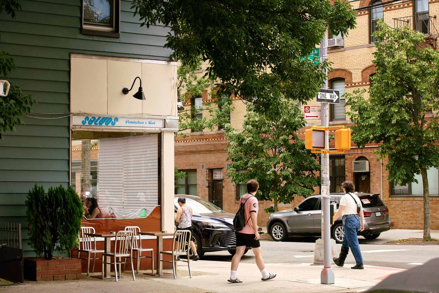 A street corner with a cafe, outdoor tables and chairs, three people walking, trees, parked cars, and brick buildings in the background on a sunny day.