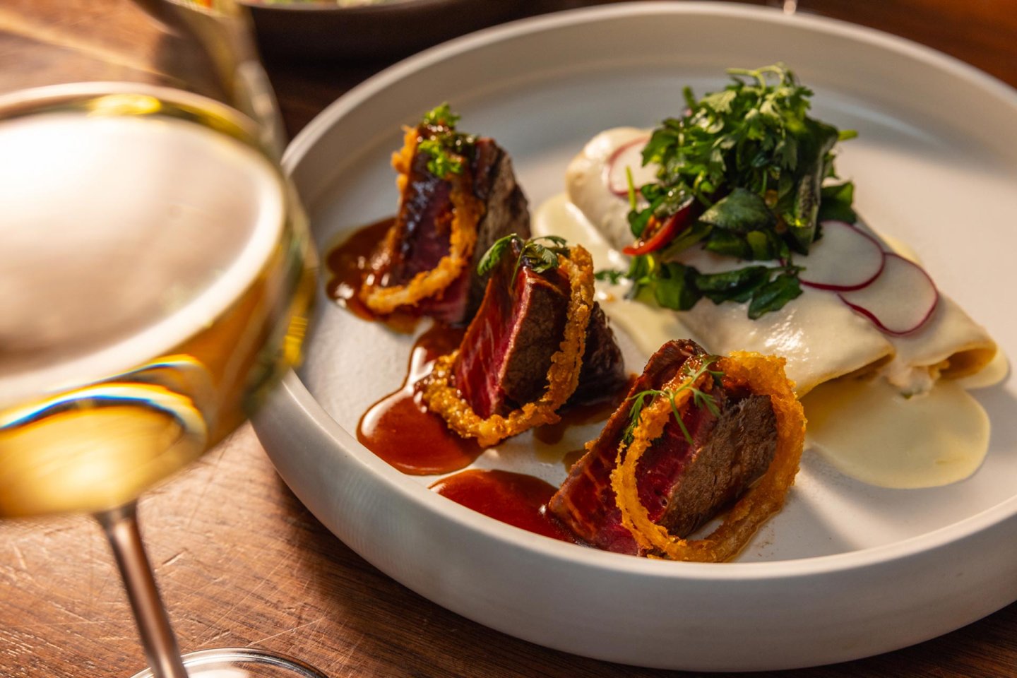A gourmet dish with seared beef in crispy rings, sauce, white fish topped with greens and radish, and a glass of white wine in the foreground on a wooden table.