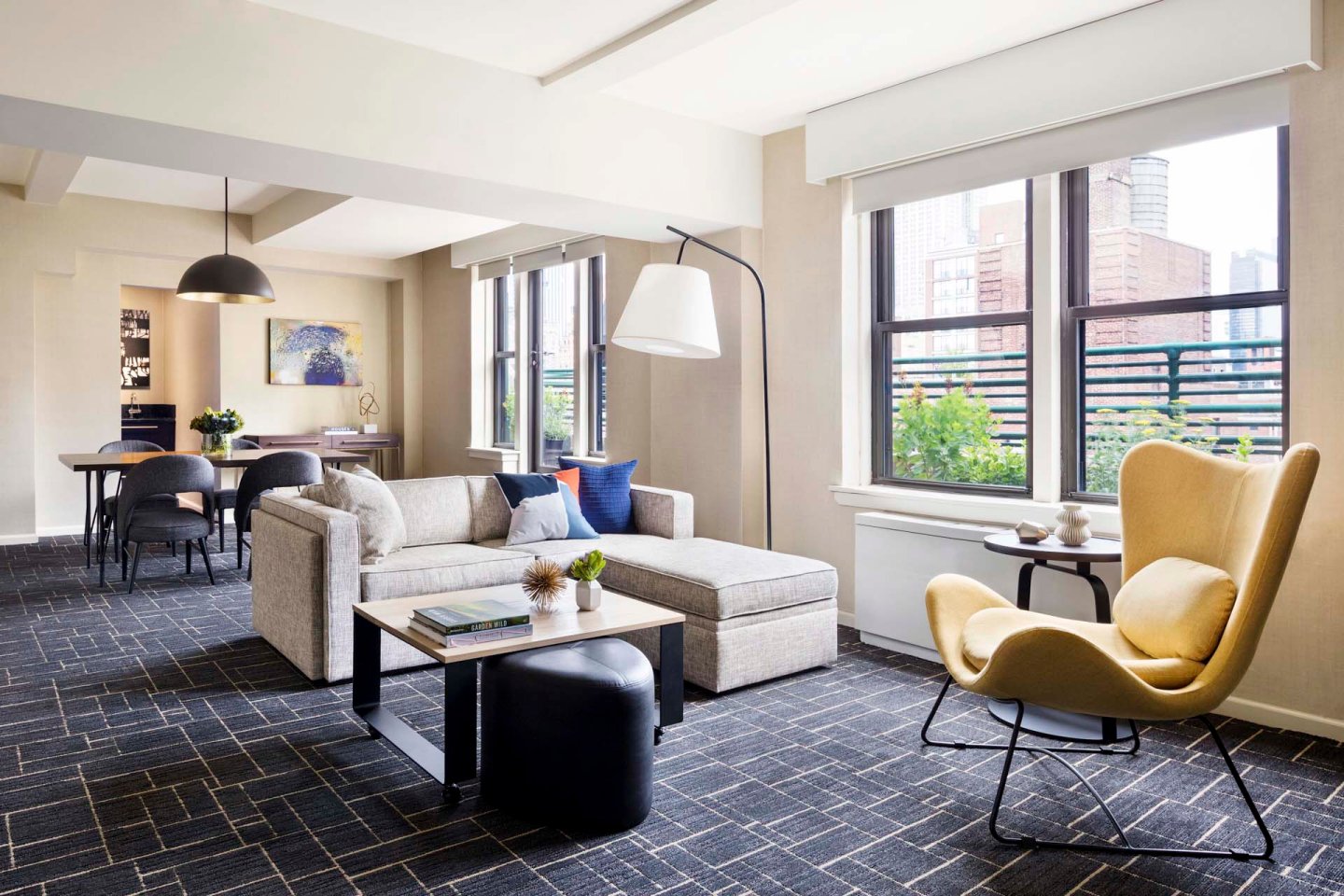 A modern living room with a grey sectional sofa, a yellow armchair, a black floor lamp, a coffee table, and large windows letting in natural light. A dining area with a table and chairs is visible in the background.