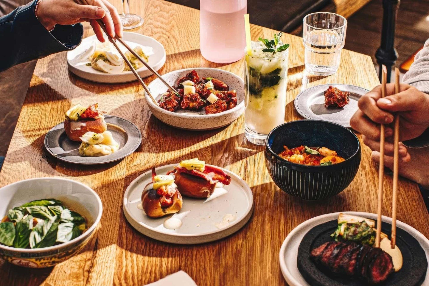 A wooden table set with assorted dishes, including sushi, small plates of appetizers, a bowl of greens, a cocktail, and water. Two people are using chopsticks to pick up food. Sunlight streams across the scene.