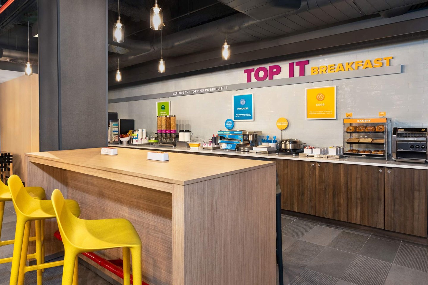 A modern hotel breakfast area with a wooden counter and bright yellow barstools, food stations along the wall, coffee machines, and signs reading “TOP IT BREAKFAST.” The space is clean and brightly lit.