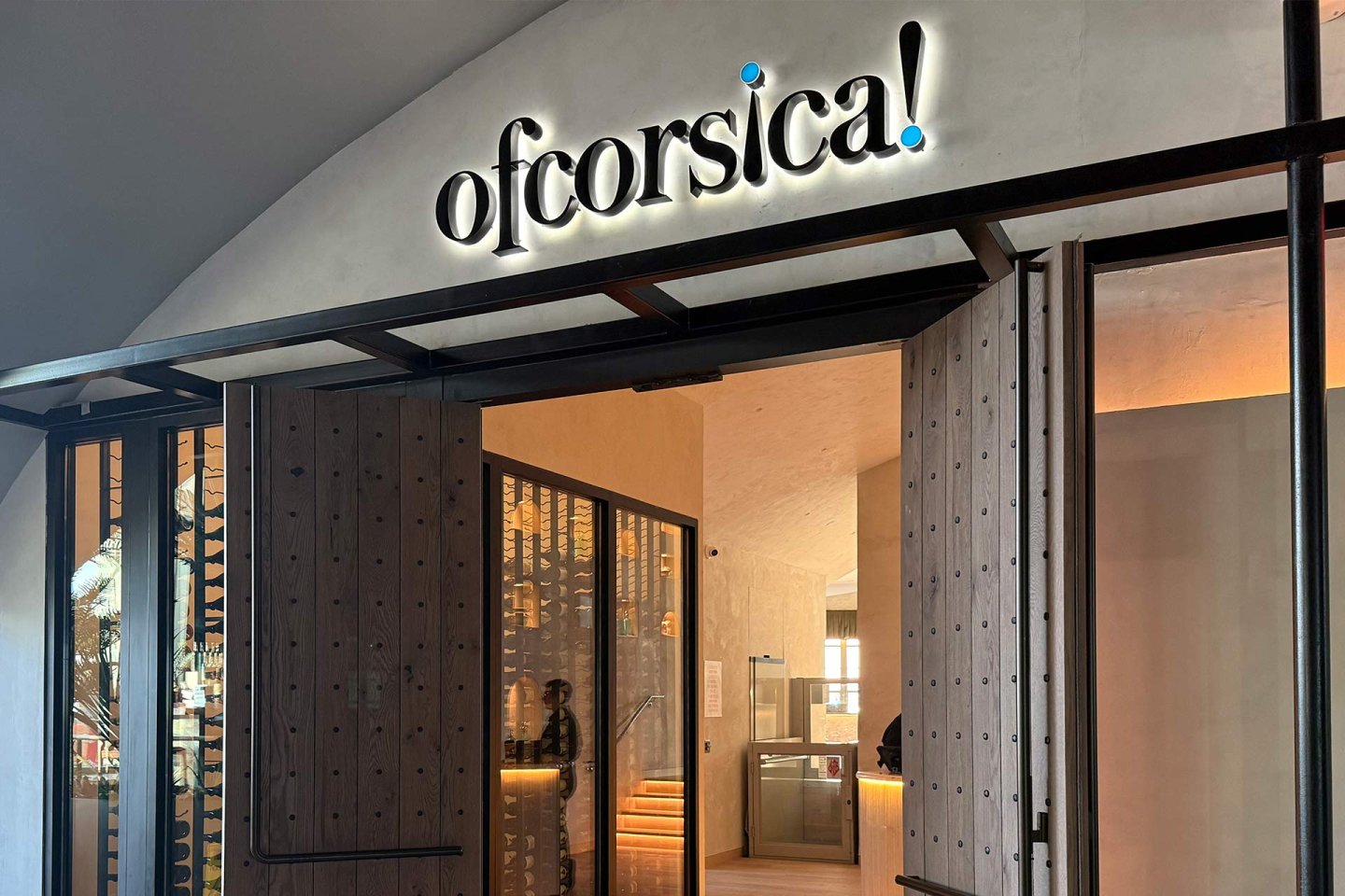 Entrance to a modern restaurant or business with a sign above the door that reads "ofcorsica!" in bold, illuminated letters. The open doors reveal a warm, inviting interior with wood and glass accents.