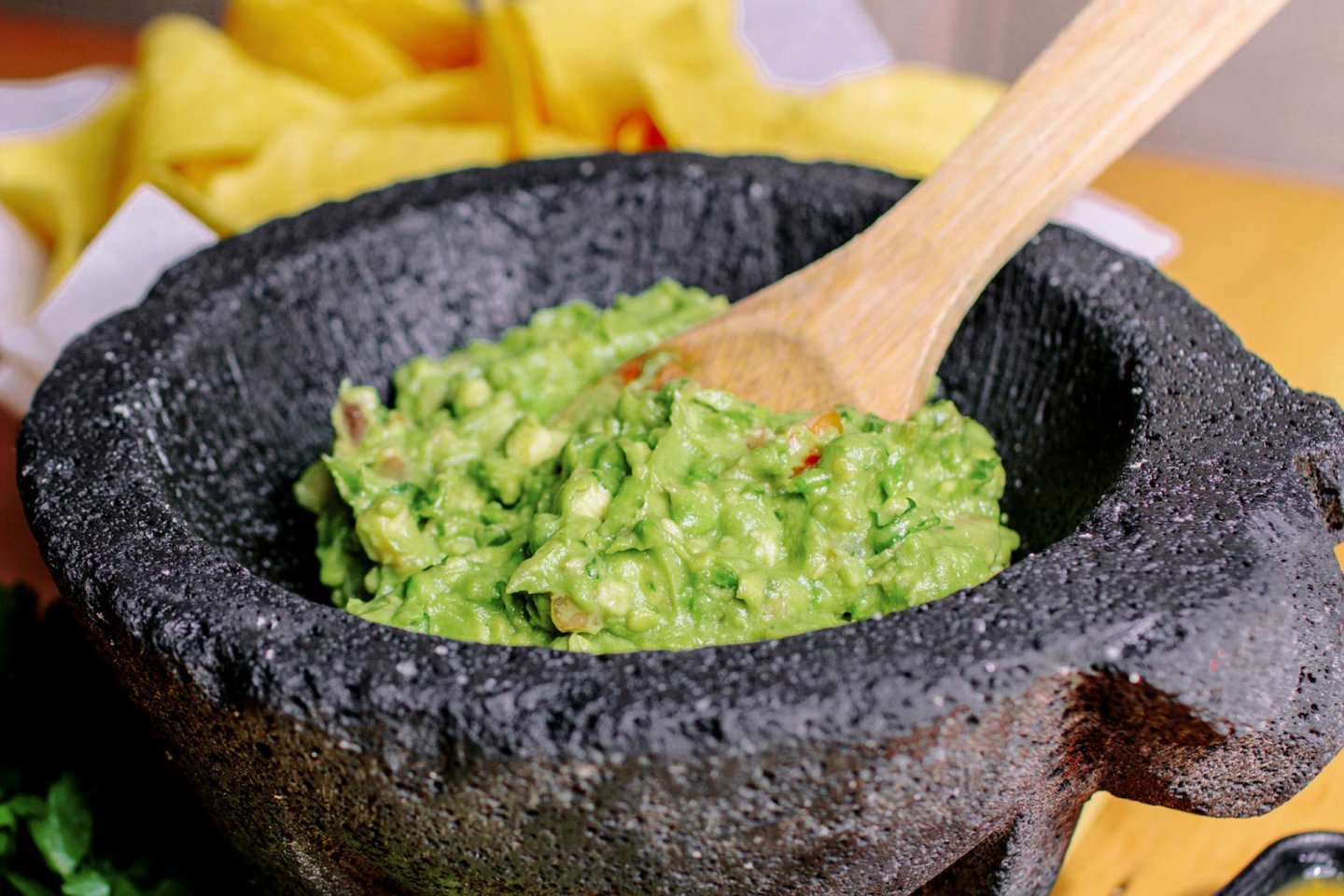 A wooden spoon stirs fresh guacamole in a traditional black stone mortar, with a yellow cloth in the background.