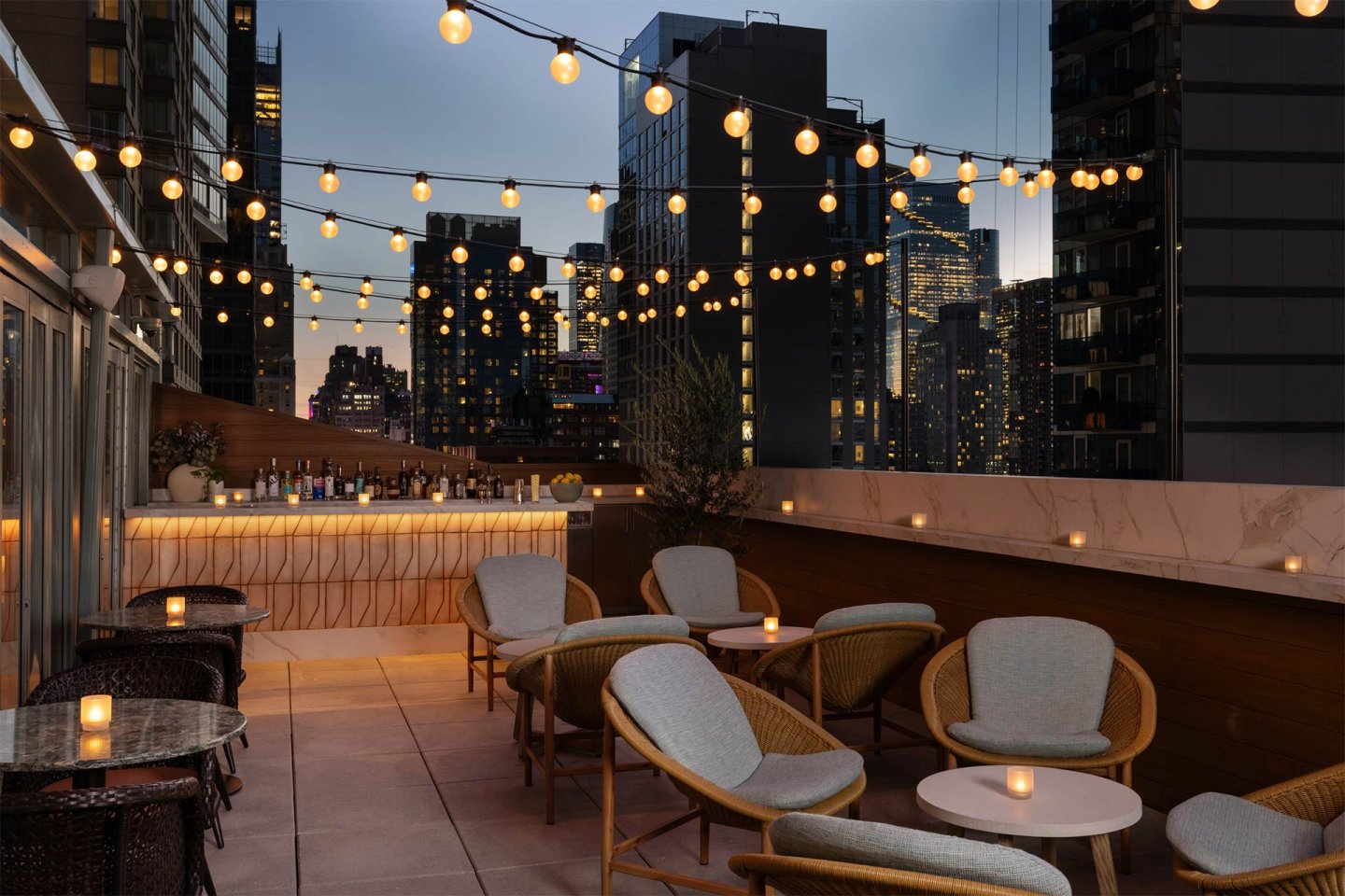 A rooftop lounge at dusk with round tables, cushioned chairs, string lights overhead, and skyscrapers in the background. Small candles are on each table, creating a cozy, inviting atmosphere.