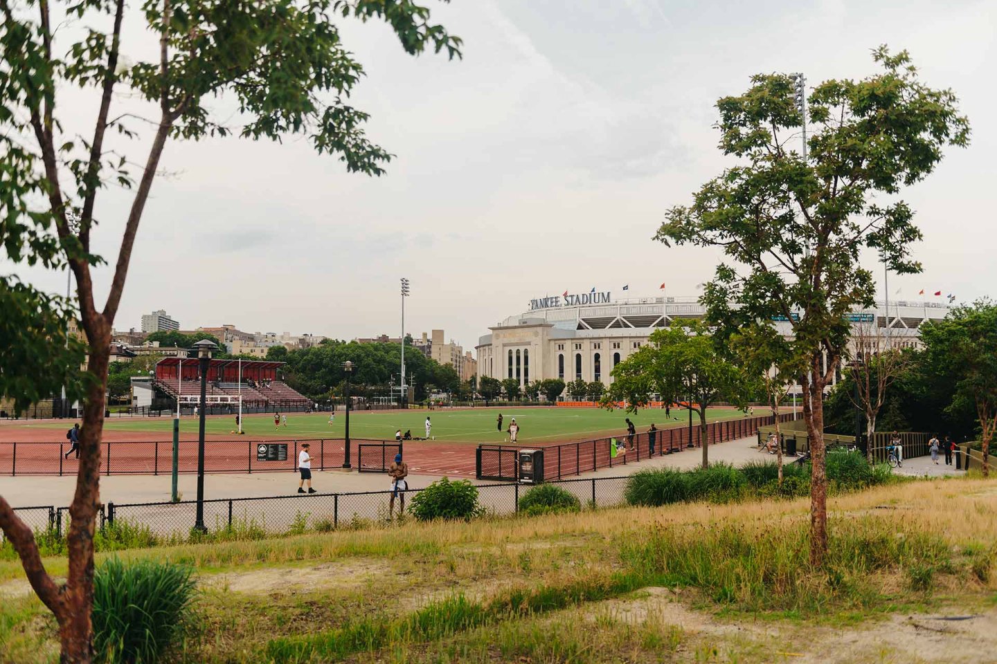 A sports field with people walking and playing, surrounded by trees and greenery, with Yankee Stadium visible in the background under a cloudy sky.
