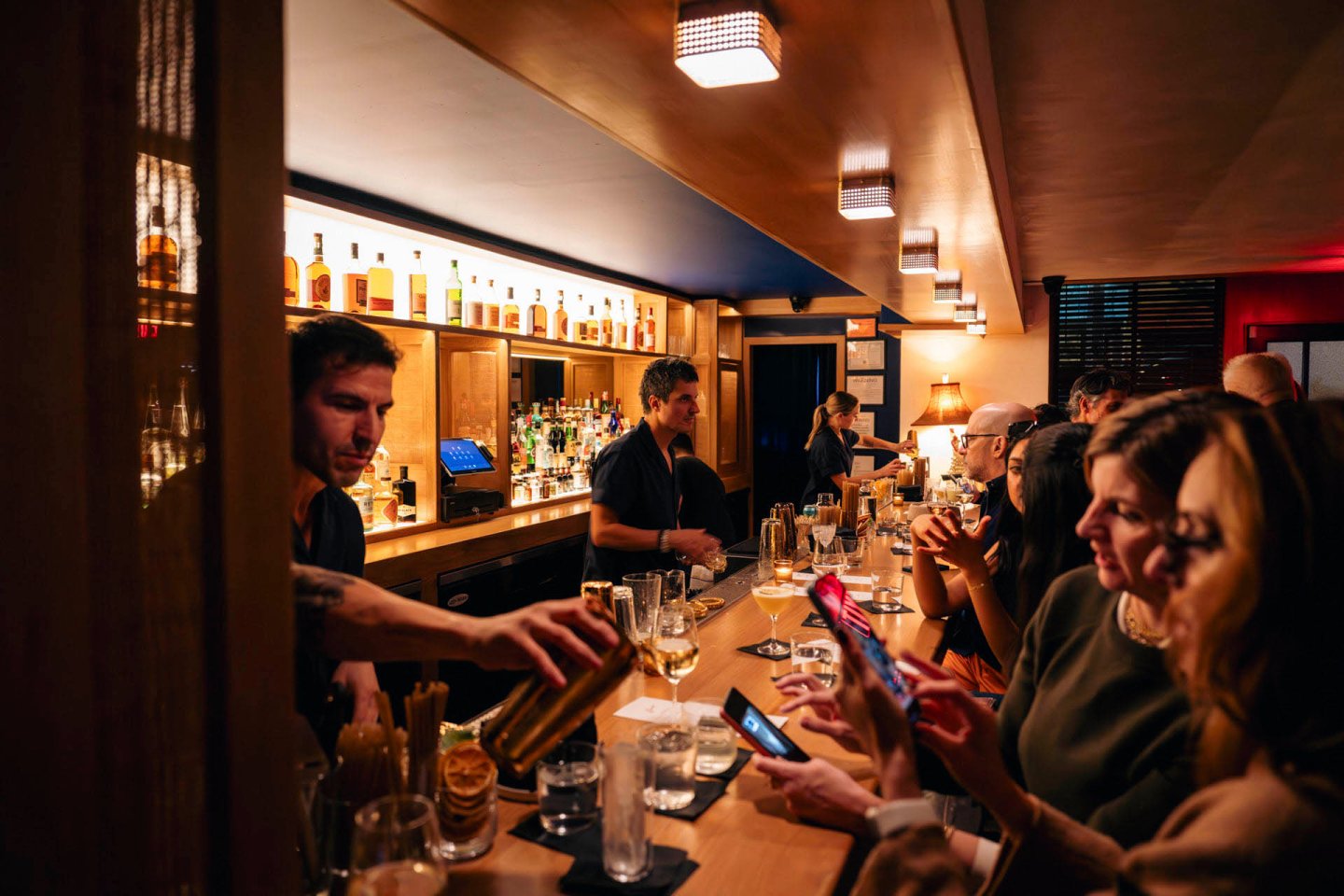 Bartender serving drinks to patrons in a dimly lit bar. Bottles of alcohol are displayed on shelves behind the bar. People are seated at the counter, some using their phones, while others chat and enjoy their drinks.