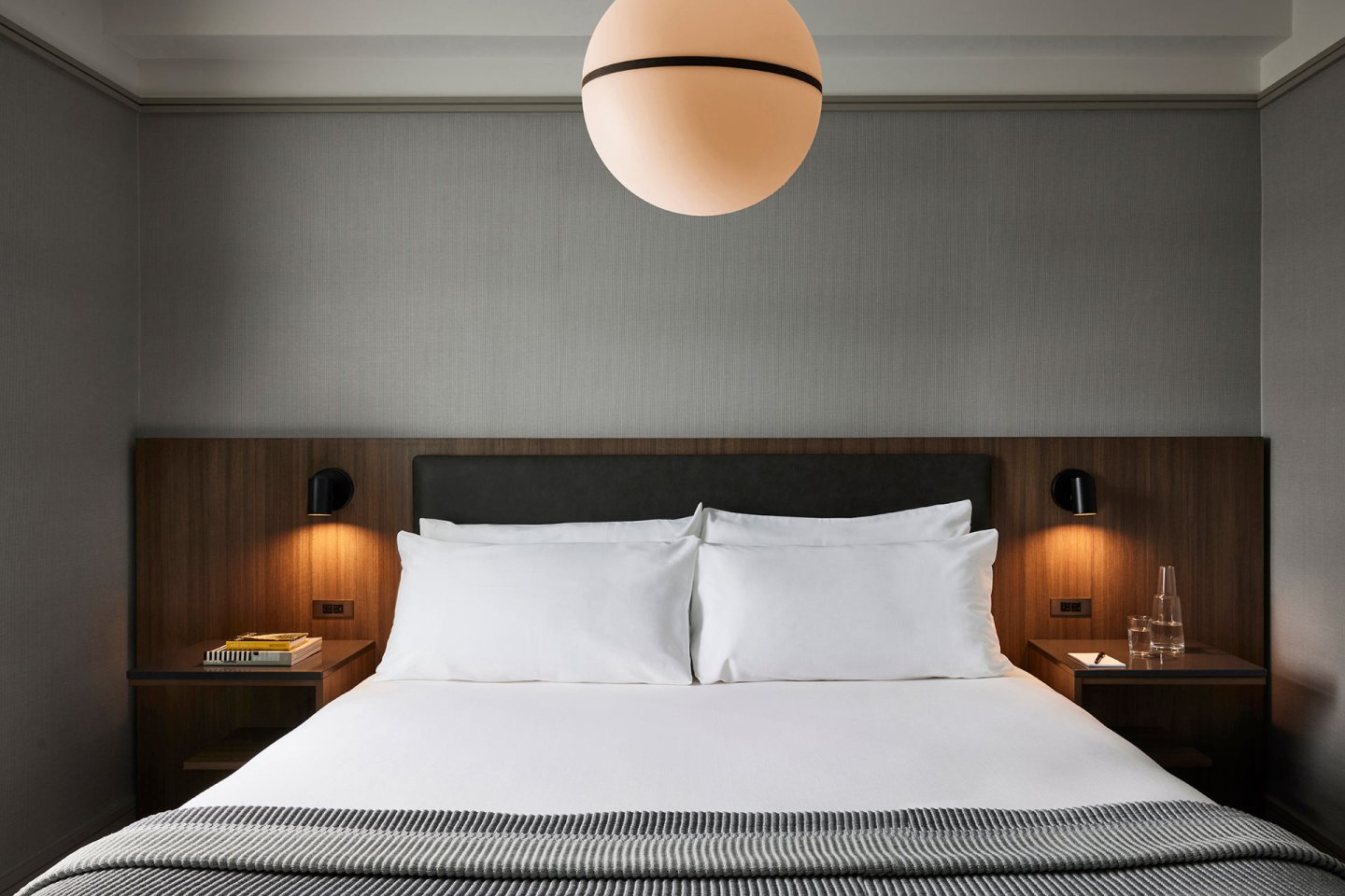 A modern hotel room with a neatly made bed, white linens, and a dark wood headboard. There are bedside tables with lamps, books, and a glass water bottle. A large round light fixture hangs from the ceiling.