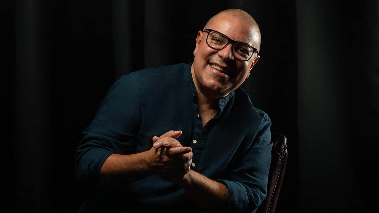 A portrait of Hector Romero, a latino man with a shaved head and glasses sitting in a chair wearing a dark blue button up shirt
