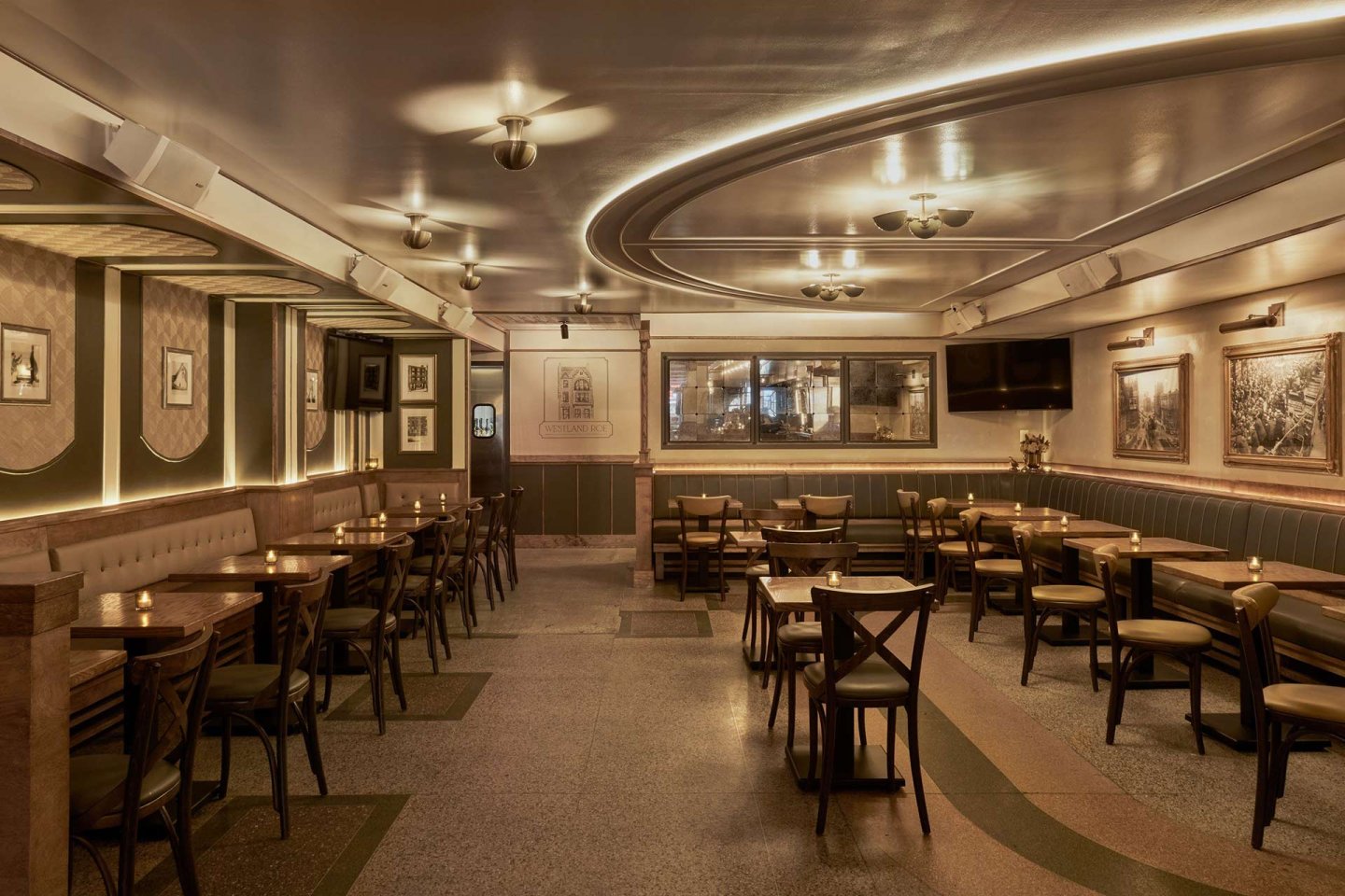 A warmly lit, empty restaurant with wooden tables and chairs, green upholstered walls, framed art, and curved ceiling lights, creating a cozy and inviting atmosphere.
