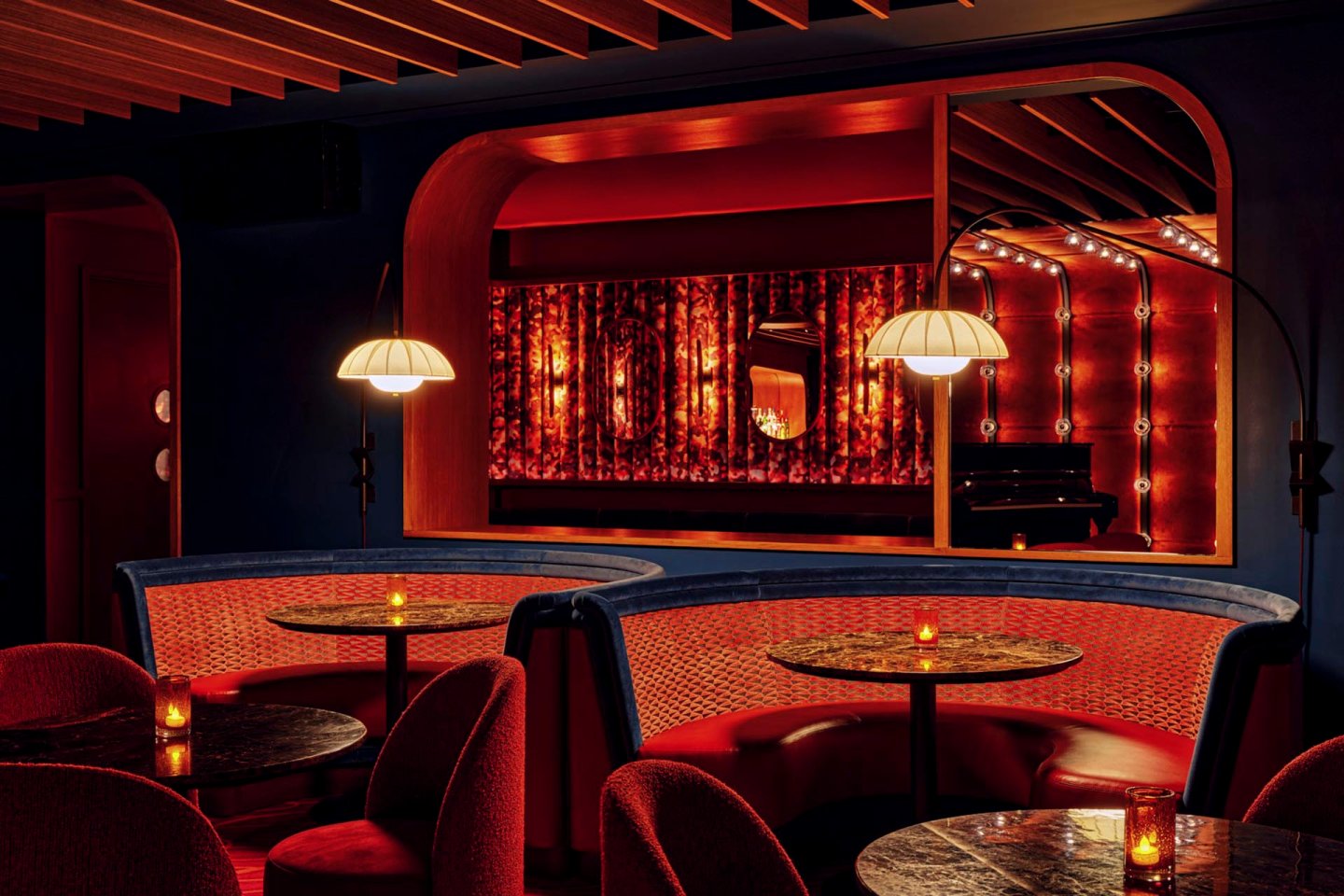 A dimly lit, elegant lounge features intimate seating with red upholstery, dark marble tables, and warm lighting from hanging lamps. The background shows a stage with a piano and glowing string lights creating a cozy ambiance.