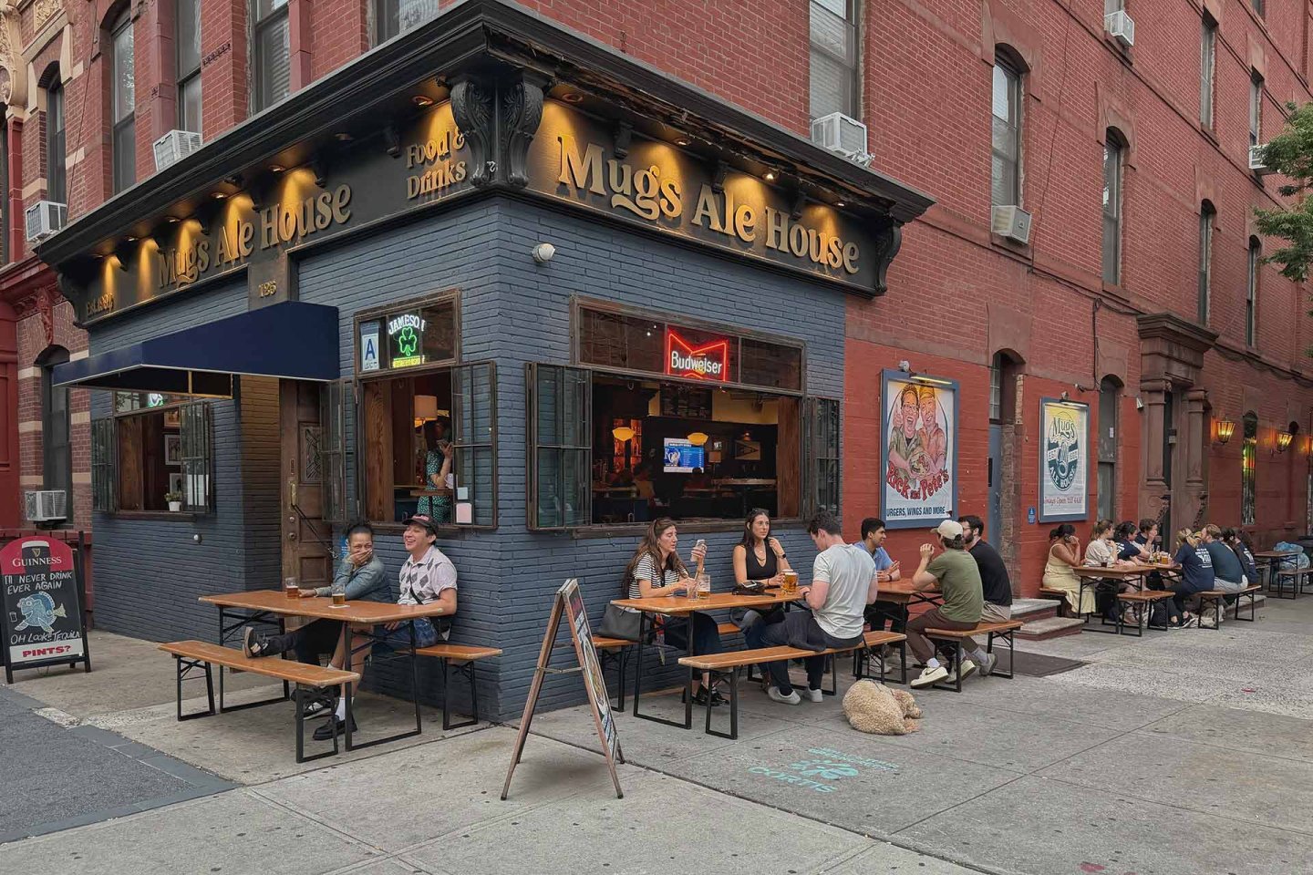 People sit at outdoor tables at Mugs Ale House, a corner bar with a dark exterior and golden signage, on a city street. Some are eating and drinking, enjoying a casual atmosphere on a cloudy day.