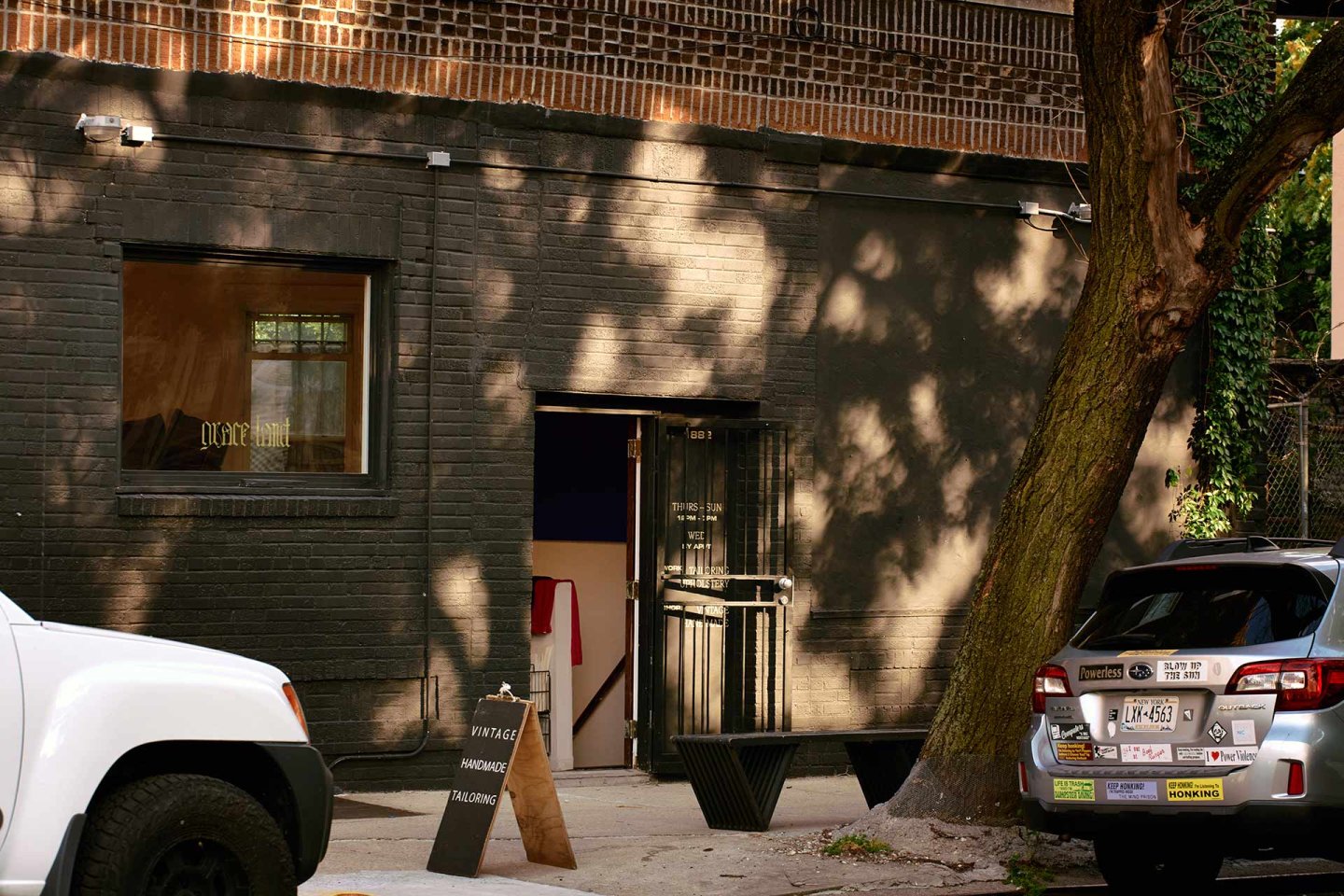 A black brick building with an open door, a white SUV, and a silver car parked outside. Sunlight filters through tree leaves, casting shadows. A chalkboard sign stands on the sidewalk near the entrance.
