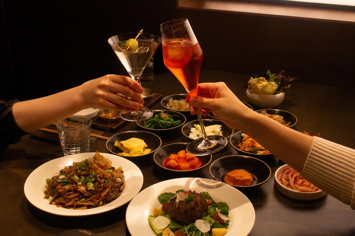 Two people clinking cocktail glasses over a table filled with various dishes, including noodles, vegetables, kimchi, salad, and a meat dish, in a warmly lit restaurant setting.