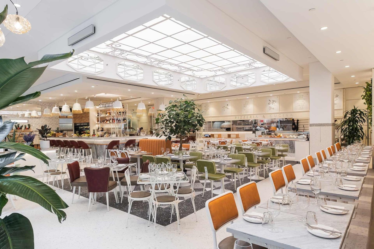 A spacious, modern restaurant with white walls, large skylights, green plants, neatly set tables, cushioned chairs, and an open kitchen in the background. The space is bright and inviting.