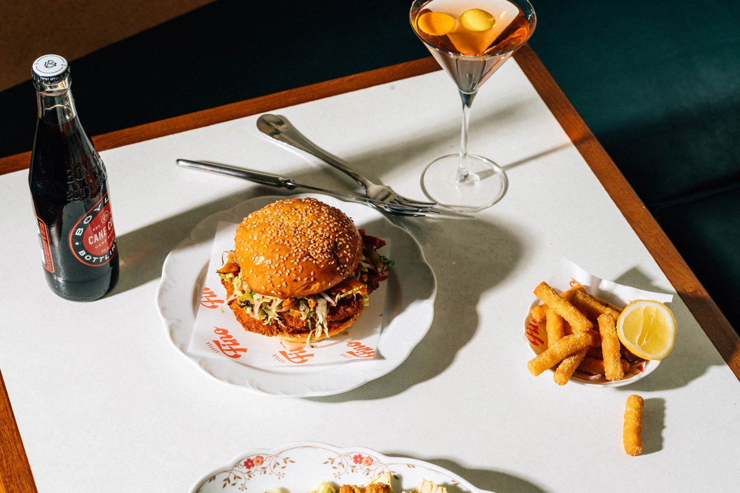 A sesame seed burger sits on a plate next to a bottle of soda, a cocktail with orange slices, and a small basket of fries with a lemon wedge, all arranged on a white table.