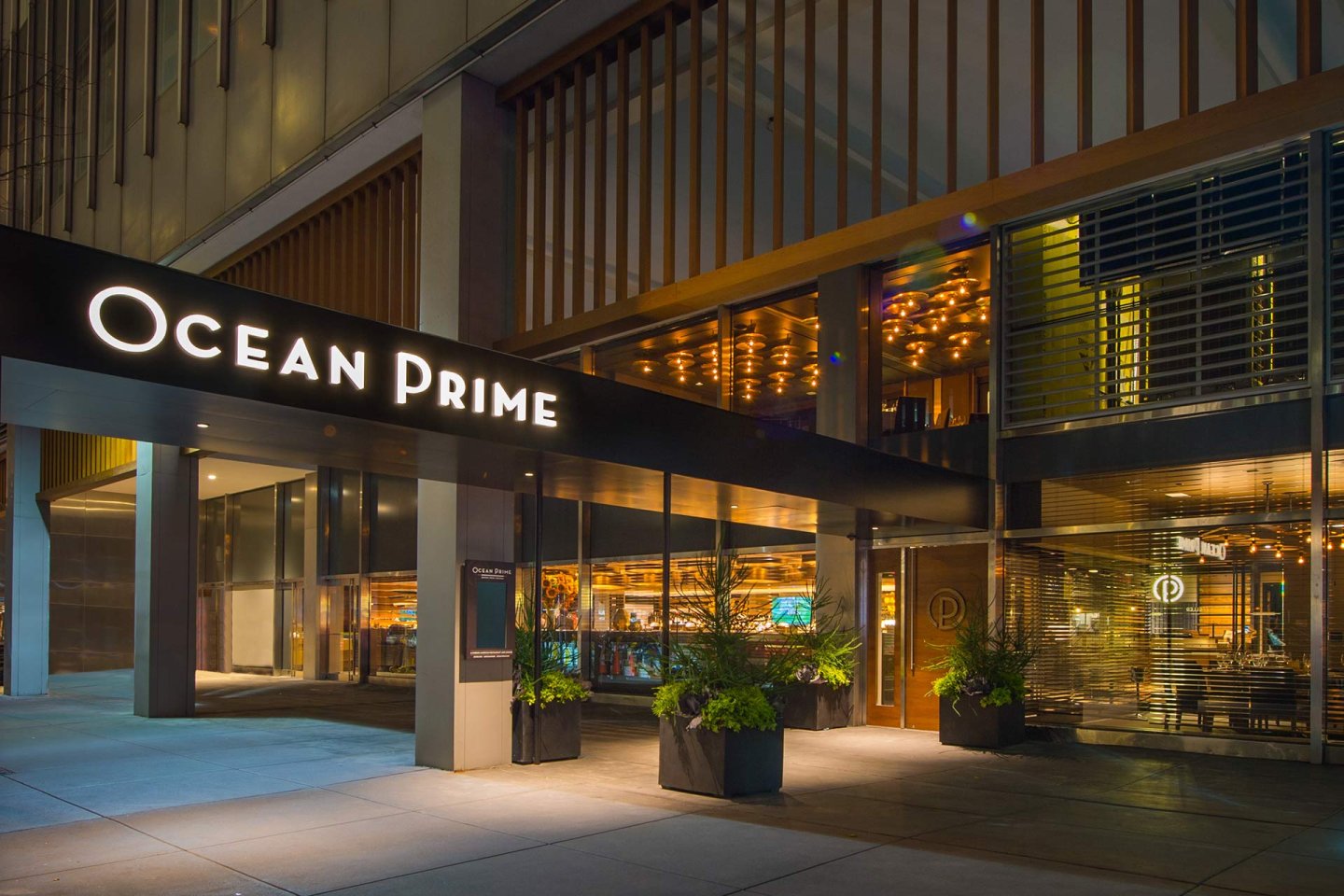Exterior view of Ocean Prime restaurant at night, with large illuminated sign, modern glass facade, potted plants at the entrance, and warm lighting visible inside.