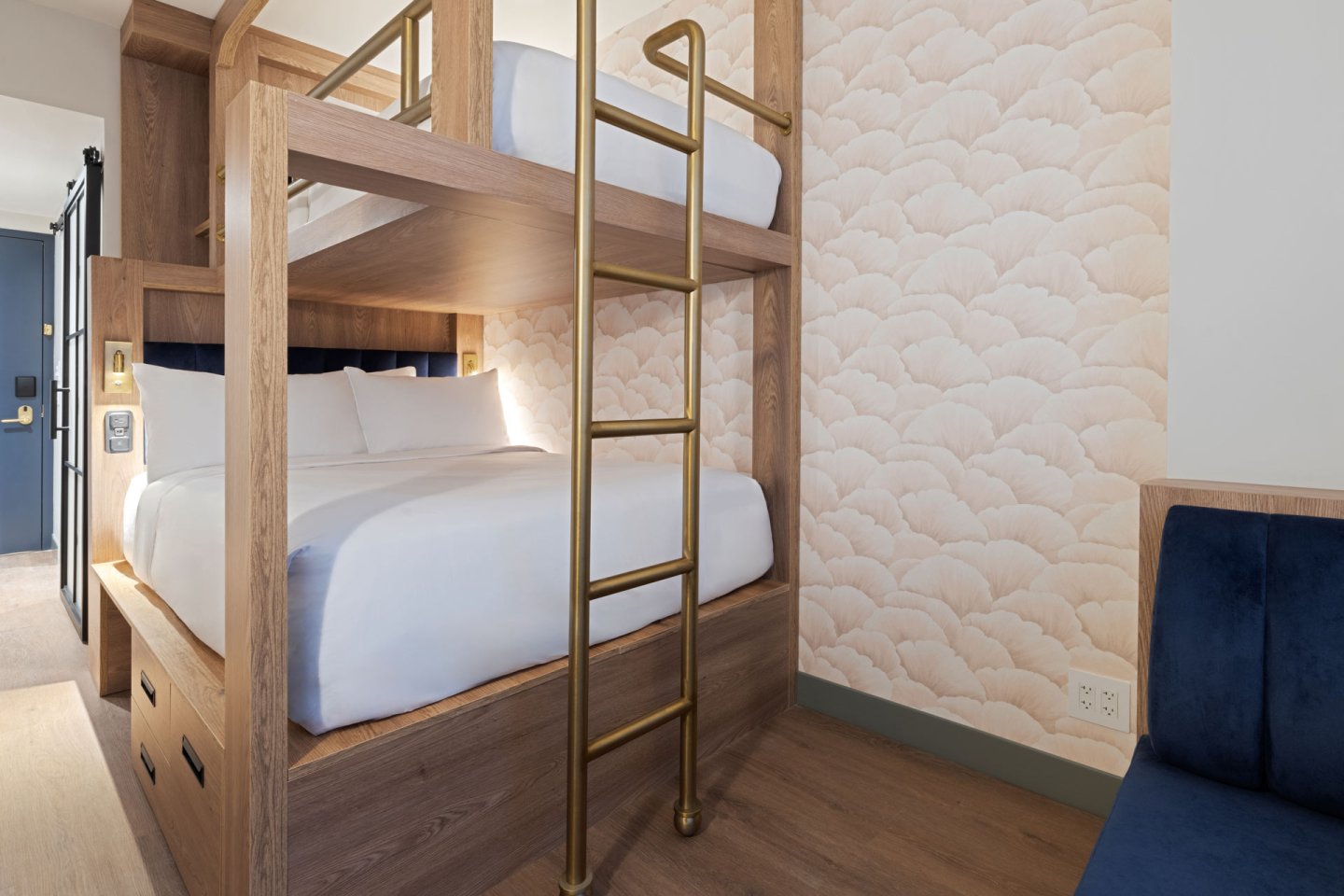 A modern hotel room with wooden bunk beds, white bedding, and a gold ladder. The walls have a soft, cloud-patterned wallpaper. A blue cushioned bench is partially visible on the right.